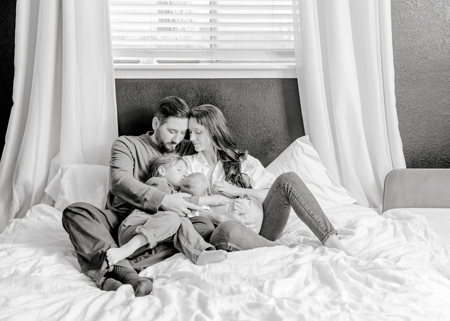 A black and white photo showing a couple cuddling and relaxing in bed with their two kids.