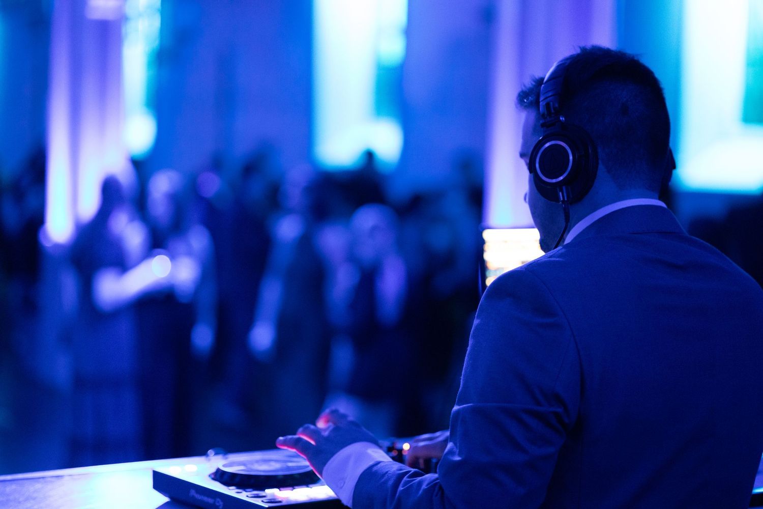 DJ wearing headphones performs at a blue-lit event, facing a crowd in an elegant venue.