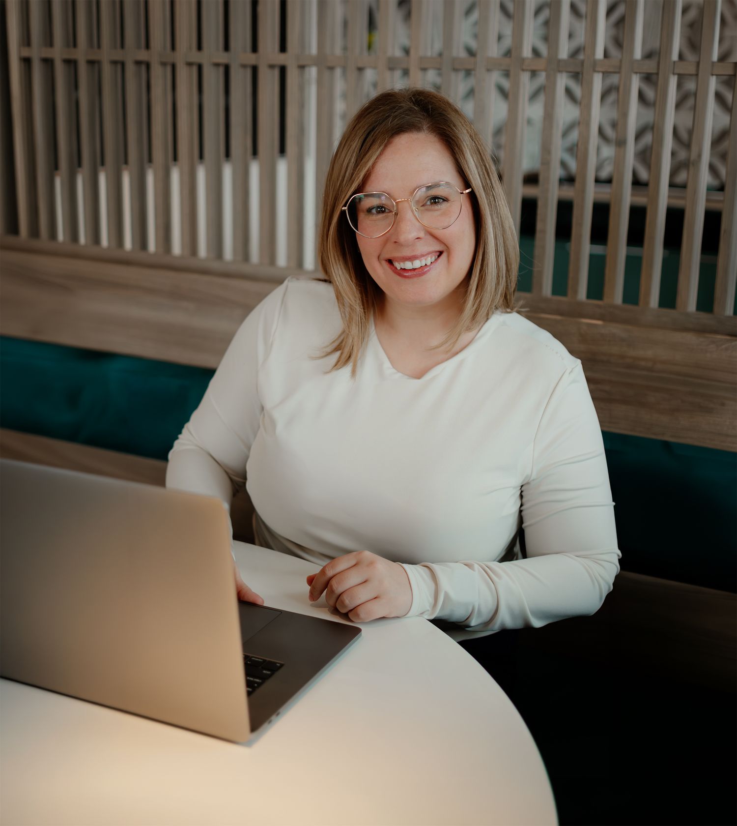 Smiling woman with glasses working on laptop at modern office desk in white top.