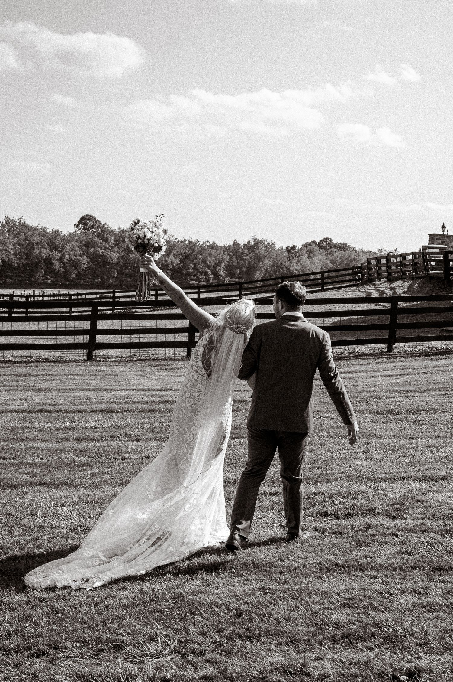 Black and white photo of bride raising bouquet joyfully while walking with groom across an open farm field.