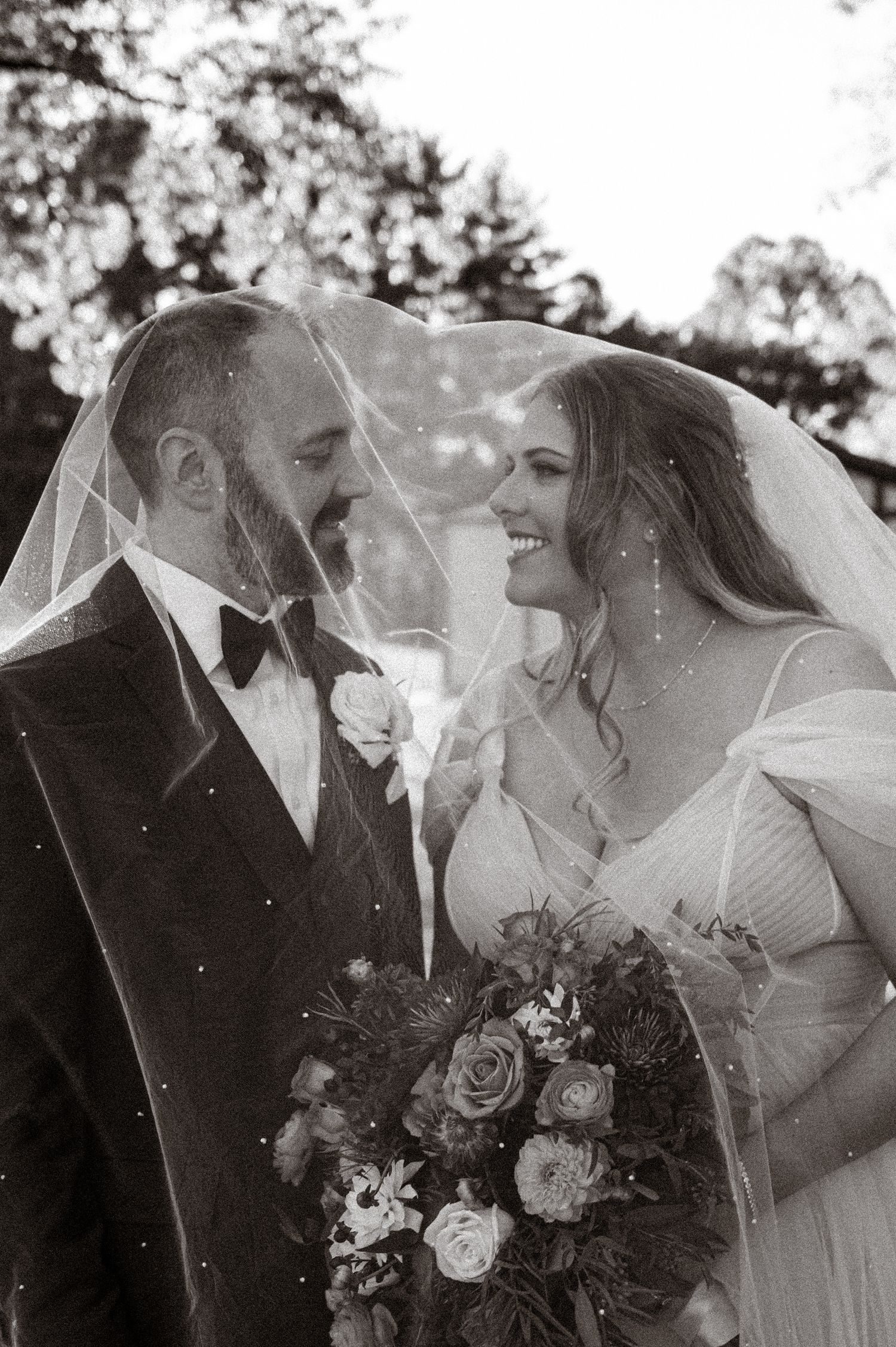 Black and white photo of bride and groom smiling under wedding veil, bride holding floral bouquet.