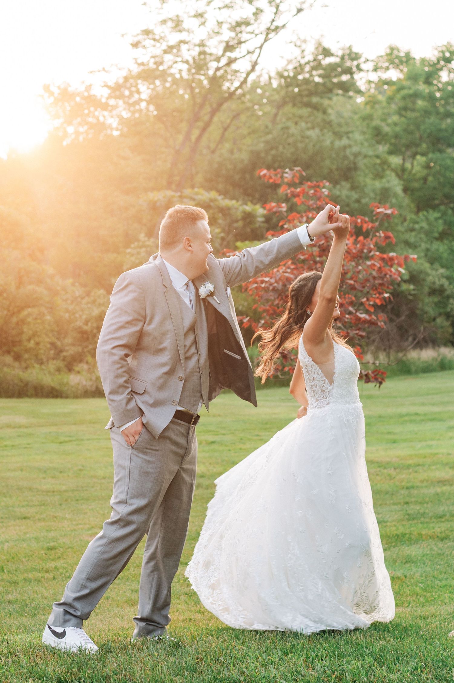 Groom twirls his bride in a lace wedding dress during a joyful outdoor dance at golden sunset.