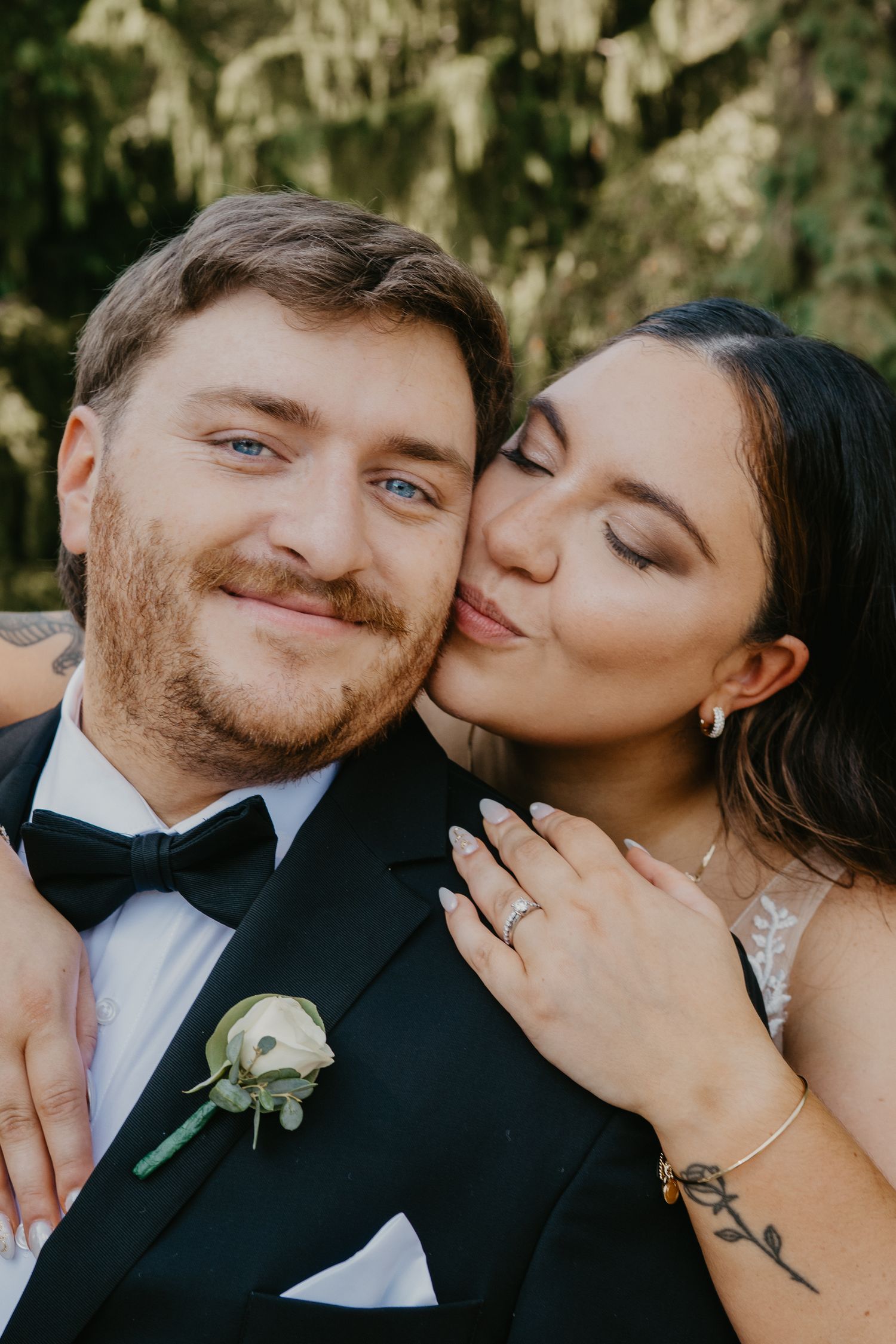 Bride kisses groom on cheek as he smiles at camera during outdoor wedding portrait.