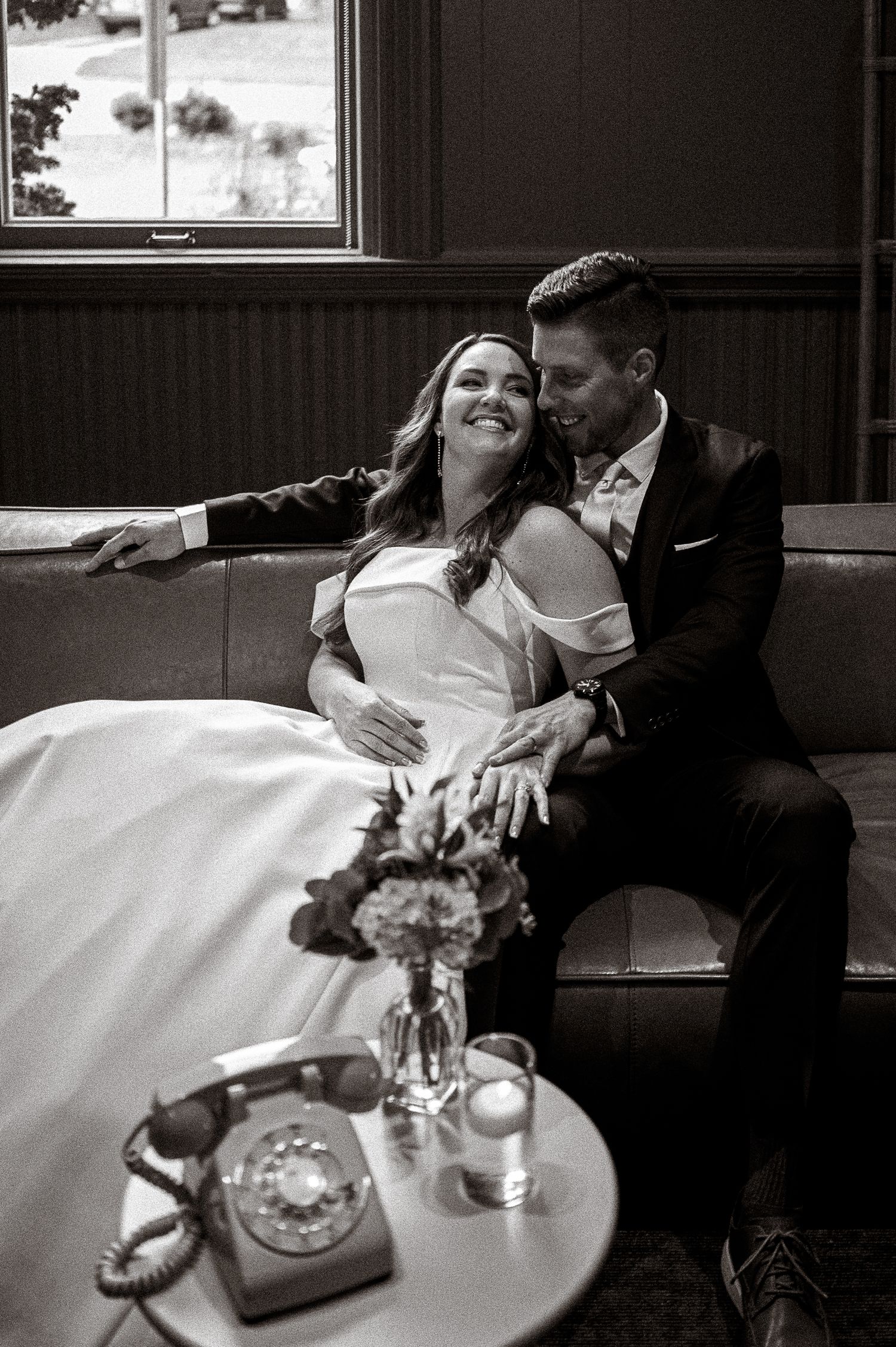 Black and white photo of bride and groom relaxing on a sofa near a vintage telephone.