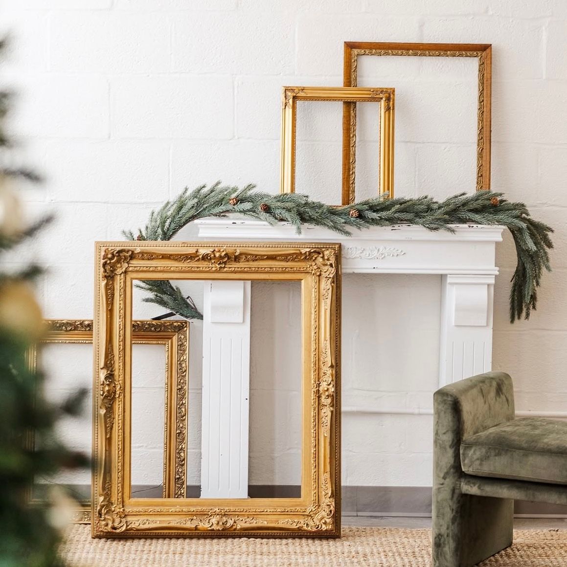 Elegant gold ornate picture frames arranged on a white fireplace mantel with pine garland and Christmas tree decor.