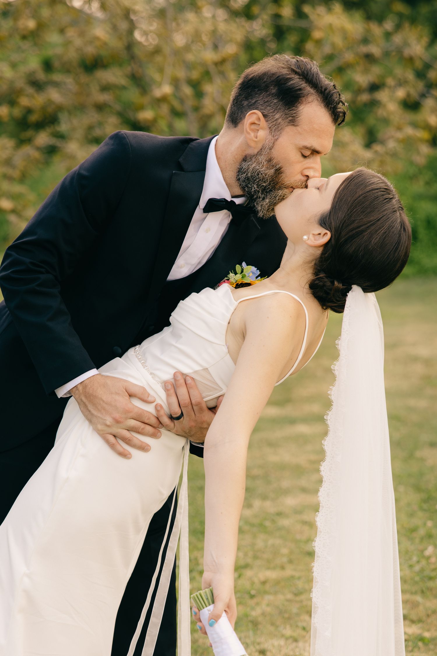 Groom dips bride for a kiss outdoors on a green lawn during wedding portraits.