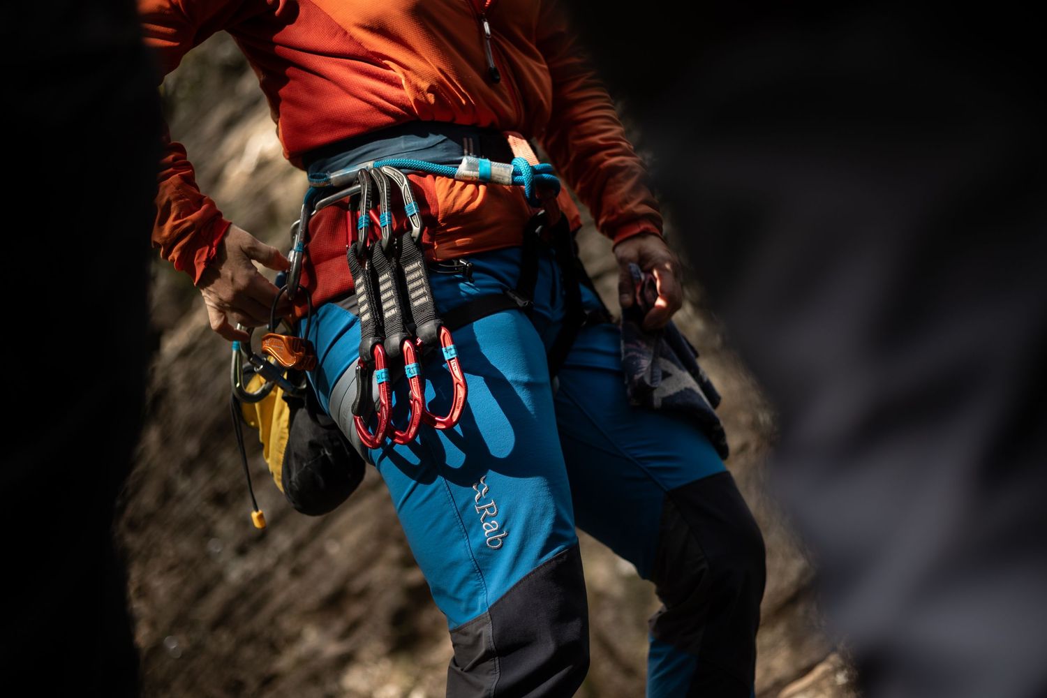 Rock climber wearing blue pants and orange jacket with carabiners and quickdraws on harness.