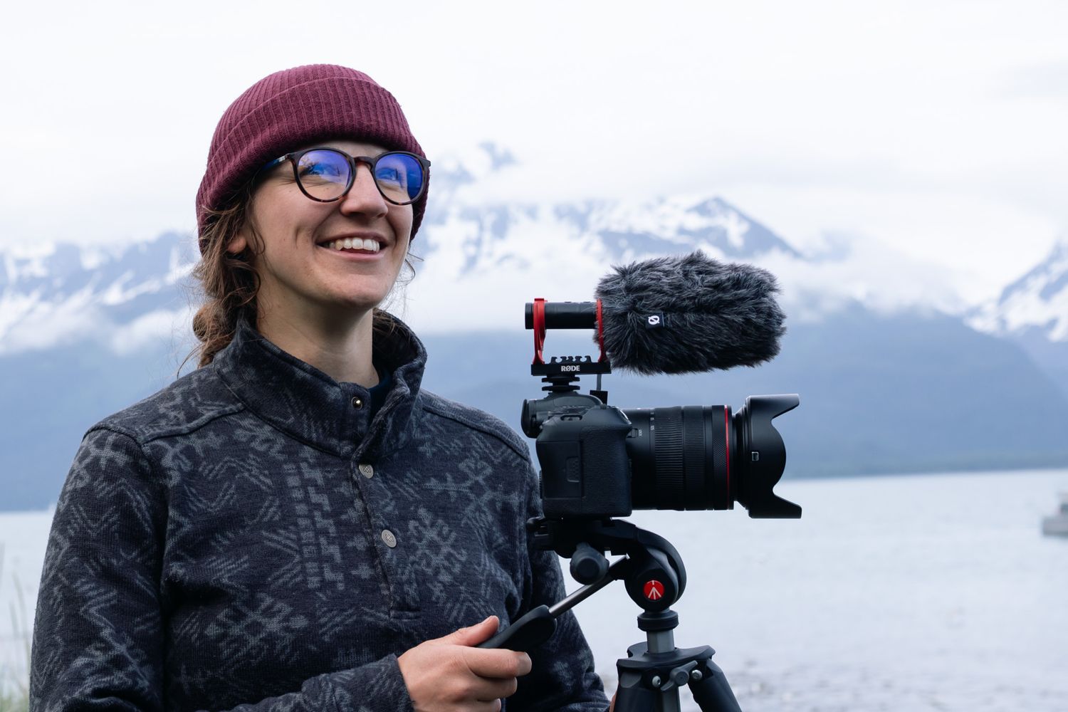 Smiling photographer with red beanie and glasses operating a camera on tripod with snowy mountains backdrop.