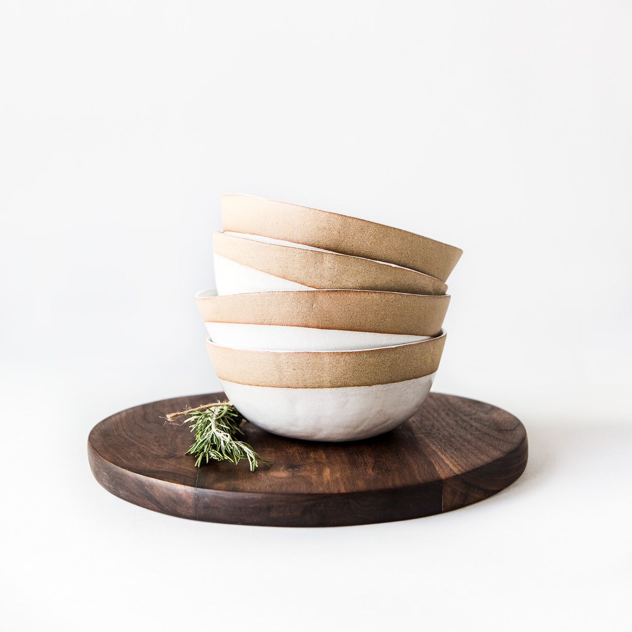 Minimalist stack of ceramic bowls on a round dark wood serving board with a sprig of greenery.