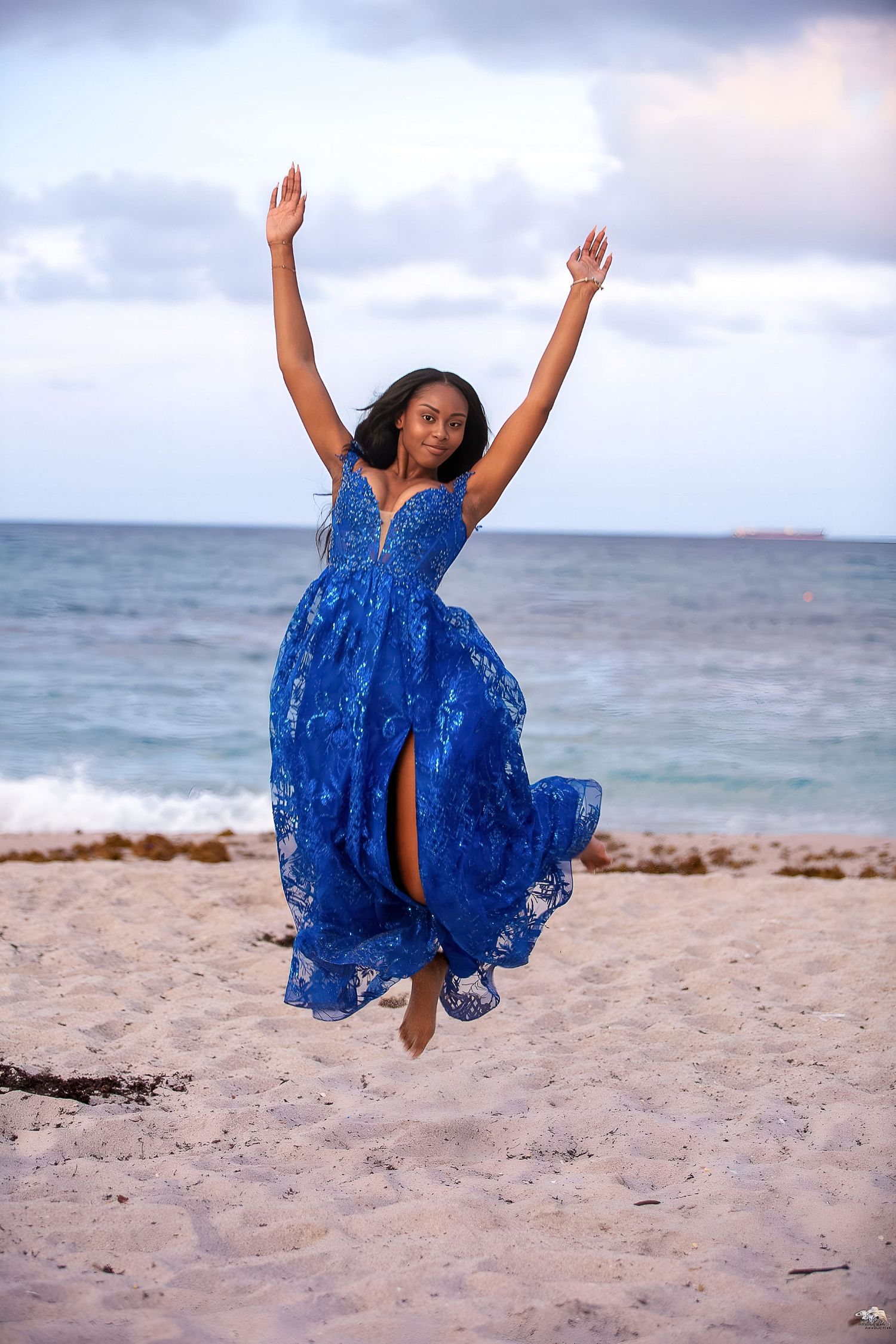 A person in a flowing blue dress jumps joyfully on a scenic beach with ocean waves in the background.