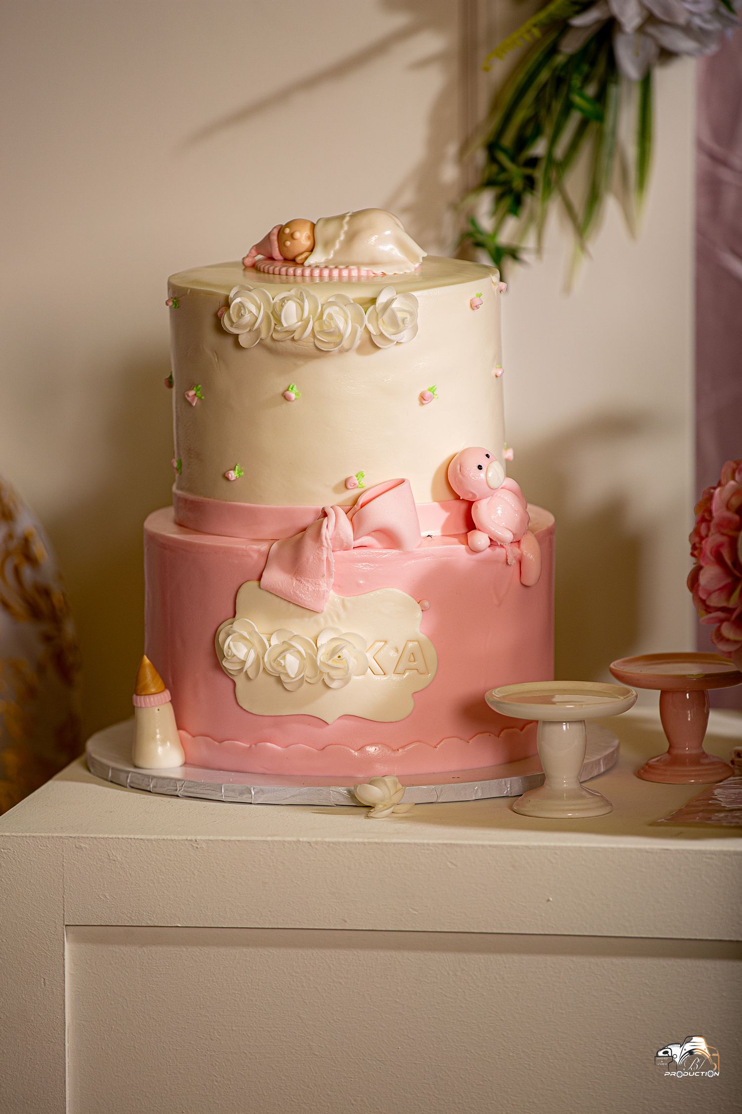 Two-tiered pink and cream birthday cake with delicate rose decorations and small animal figurines on a white display stand.