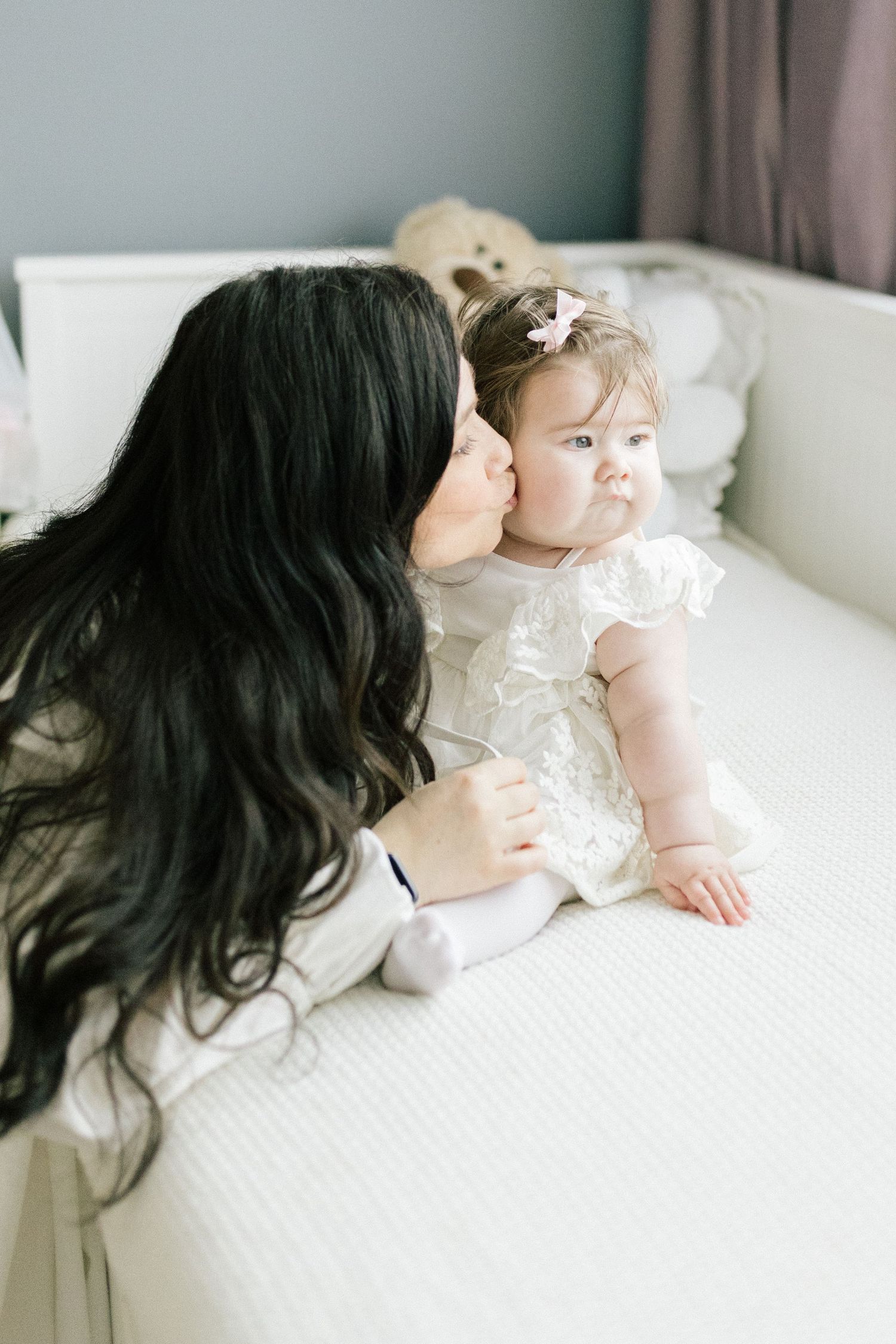 A dark-haired mother gently kisses her baby girl in a white dress on a white bed.