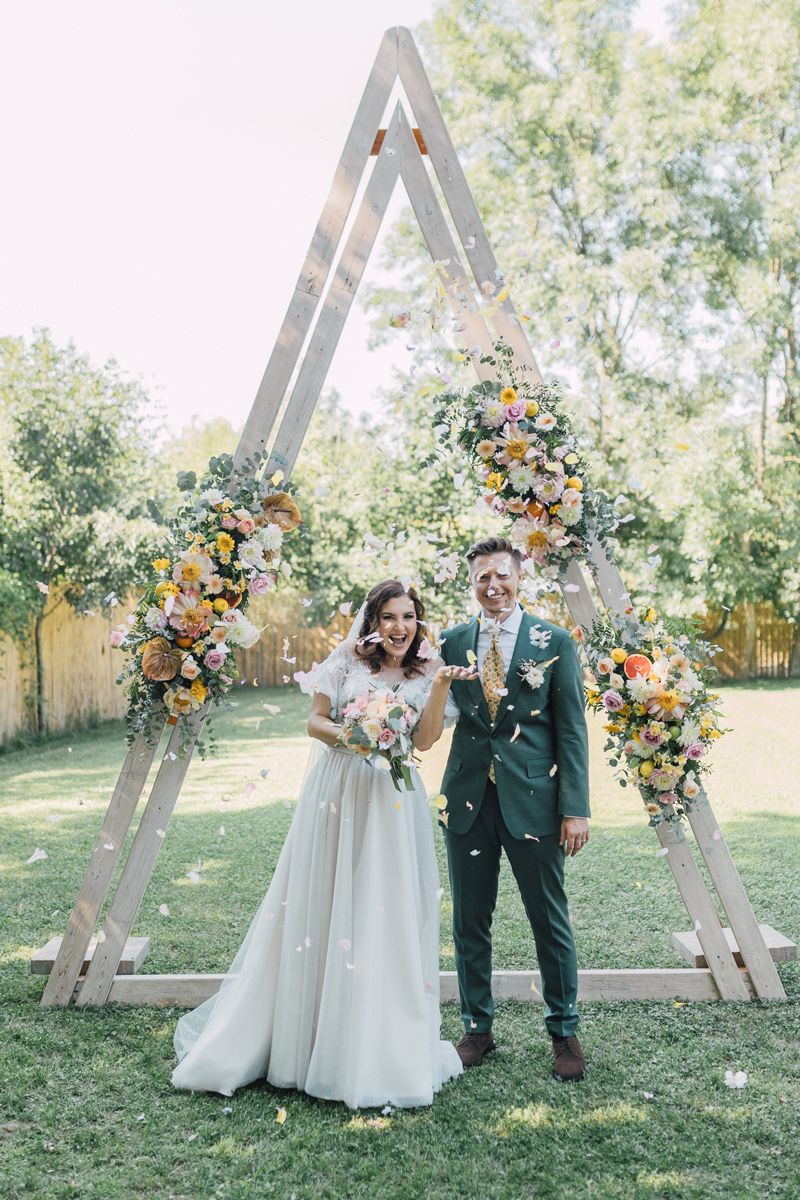 Wedding party poses in front of rustic wooden triangle arch decorated with peach and orange floral arrangements.