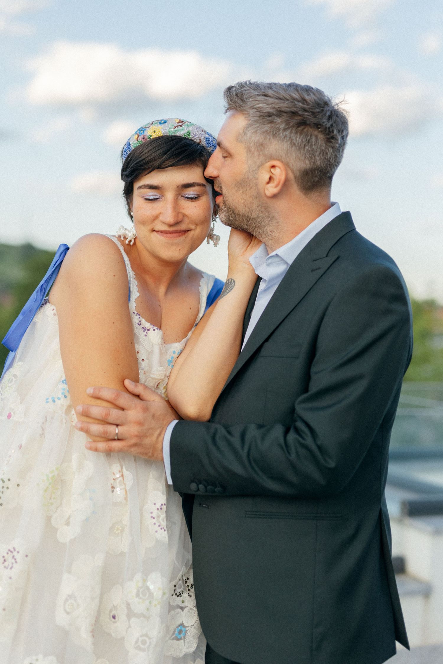 A groom in a dark suit tenderly kisses the cheek of a smiling bride in a floral white dress and floral headband outdoors.