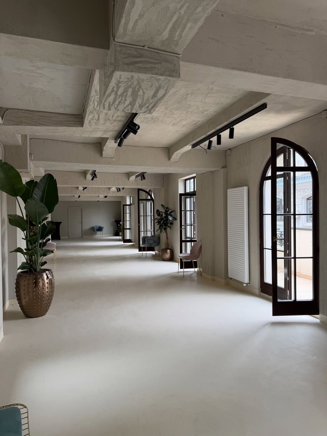 Spacious event hall with exposed concrete beams, arched black-framed windows, track lighting, potted plants and a polished light floor.