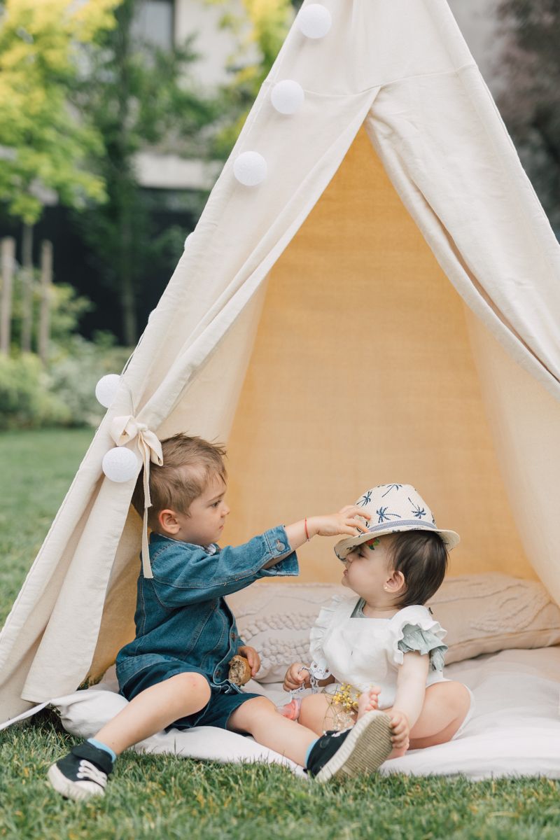 Two toddlers playing together inside a cream canvas teepee tent with pom-poms in a garden.