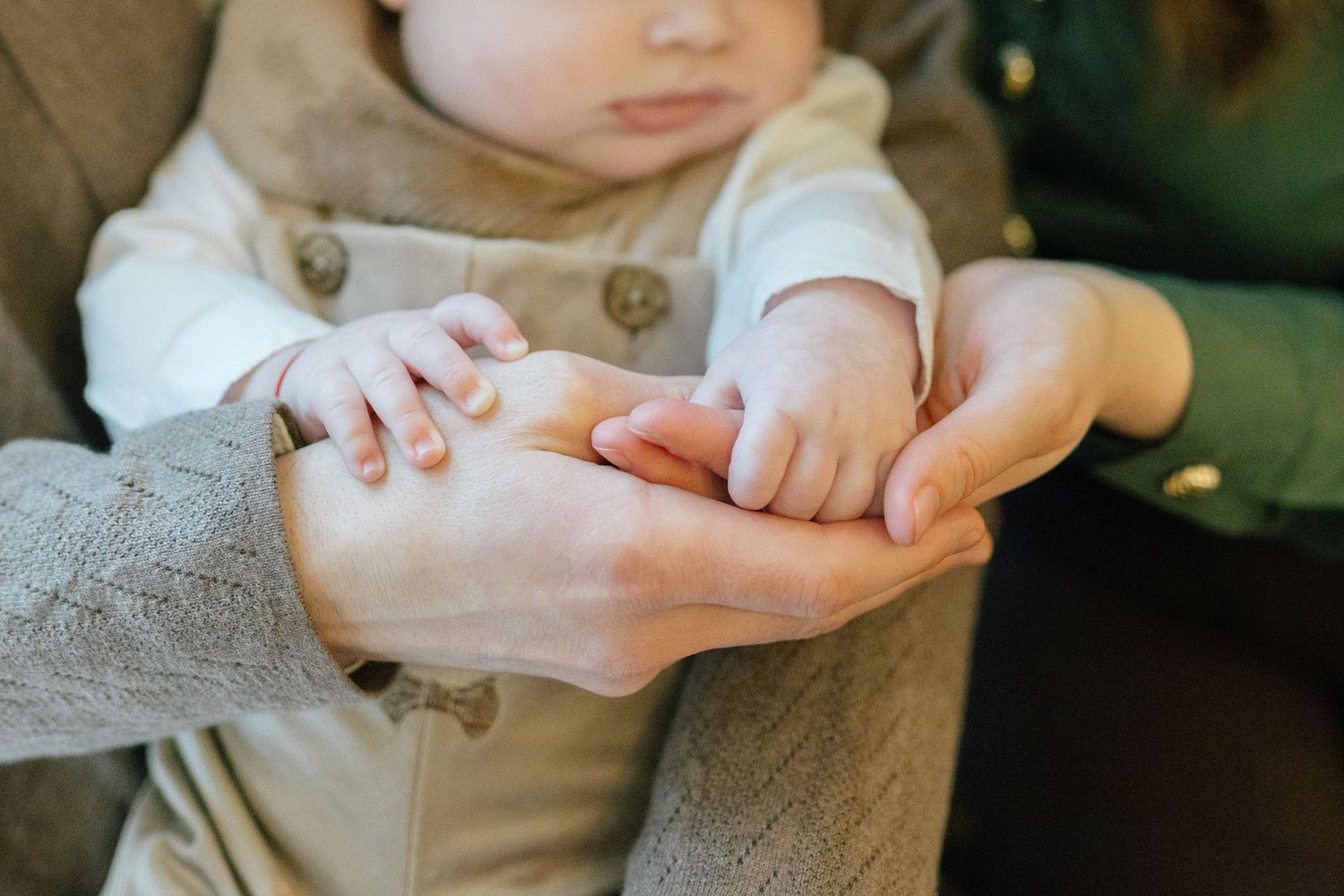 Close-up of an adult's hands gently cradling a baby's tiny hands, with the infant in a cream outfit with buttons.
