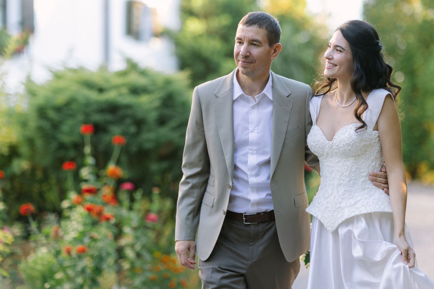 Smiling bride in a white lace gown walks arm in arm with a groom in a gray blazer past blooming red flowers in a sunny garden.