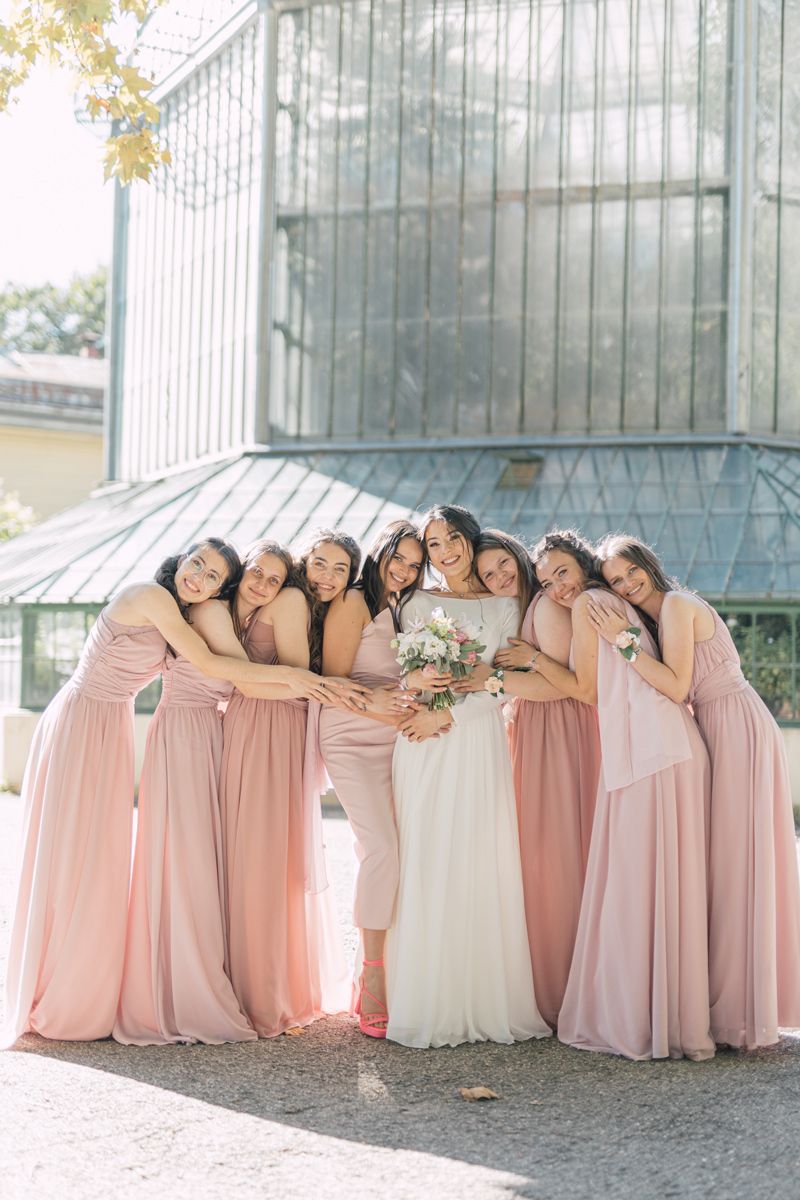 Bridesmaids in soft pink and blush dresses pose together in a creative leaning formation outside a building.