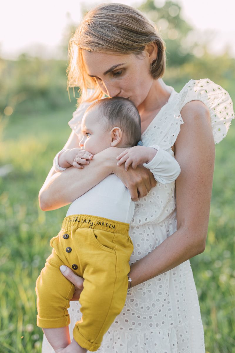 A mother in a white dress tenderly kisses her baby wearing yellow pants in a sunlit green field.