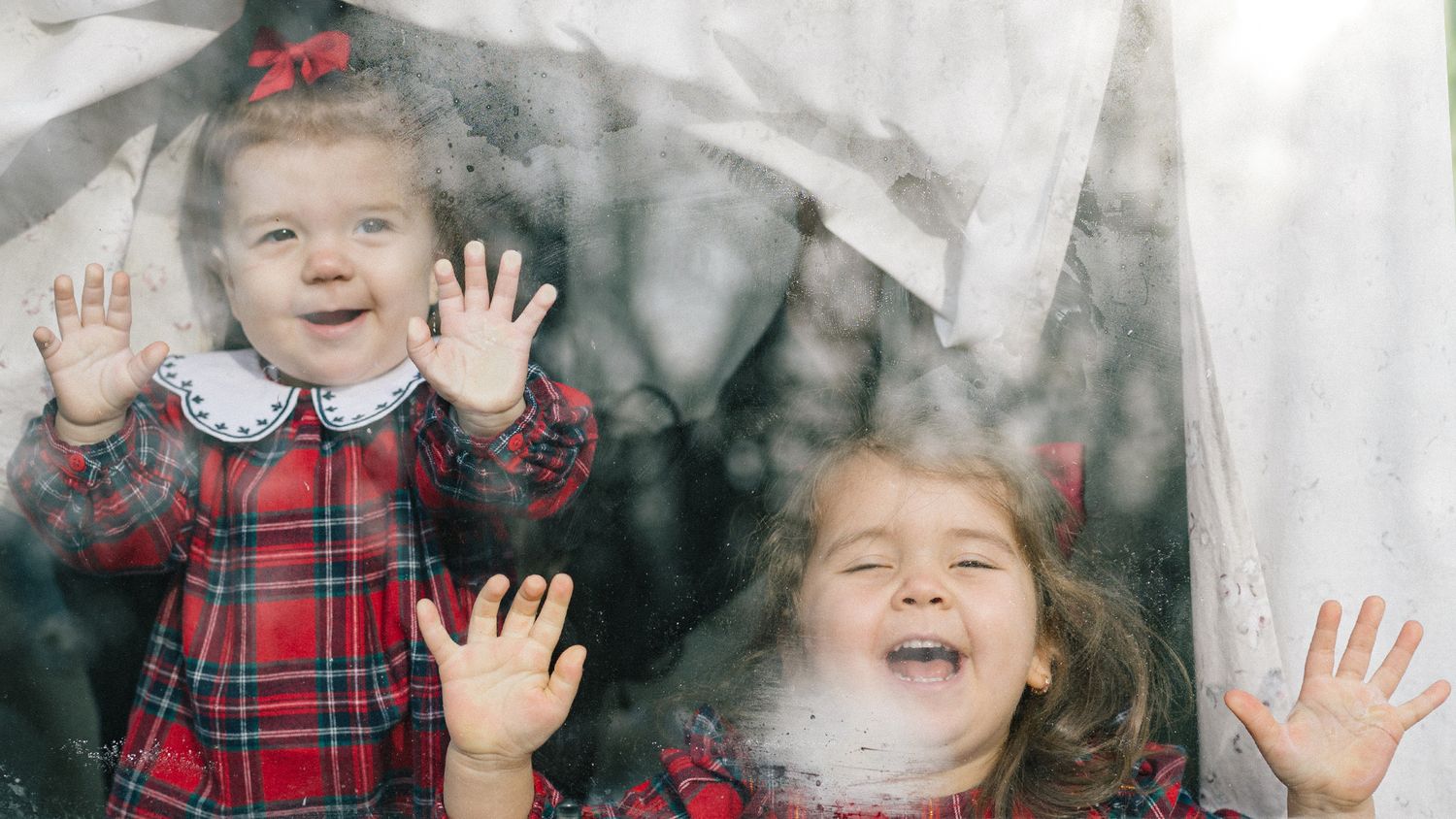 Two smiling toddler girls in red plaid dresses press their hands against a rain-speckled window with curtains behind them.