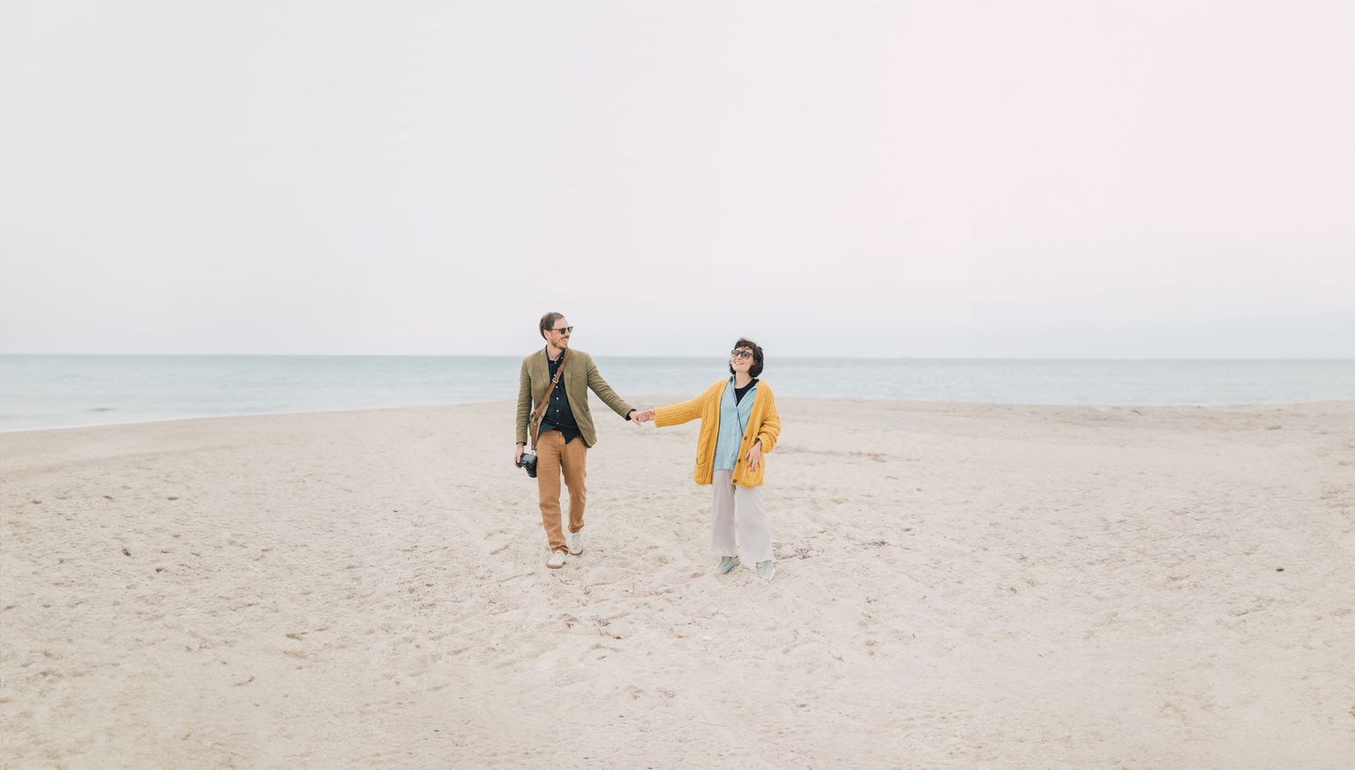 Couple in casual jackets holding hands while walking along a wide, empty sandy beach beside calm water under an overcast sky.