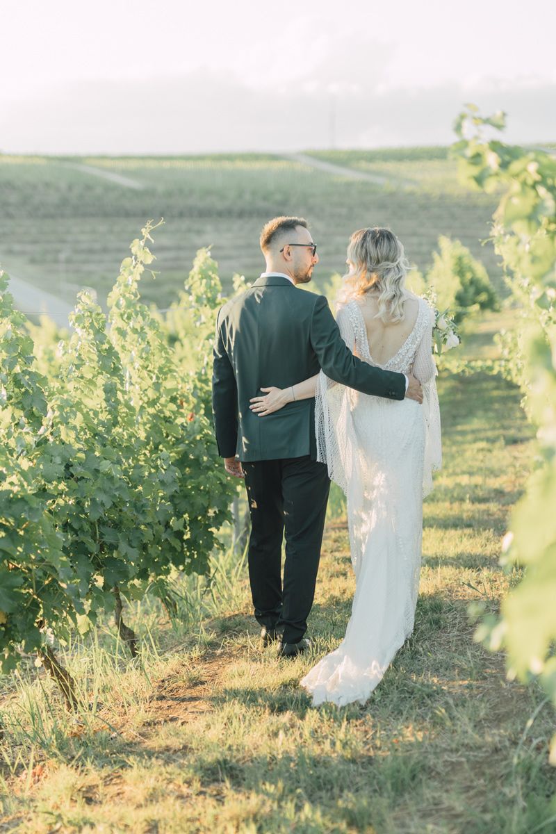 A couple in wedding attire walks hand in hand through a sunlit vineyard during golden hour.