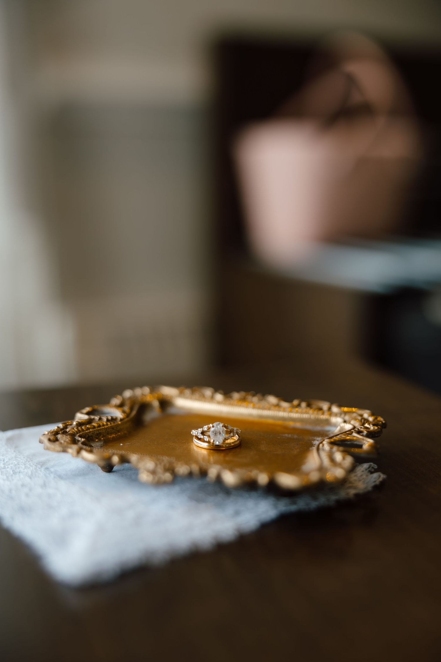 Gold ornate tray with diamond ring on white cloth atop dark wooden table, blurred background.