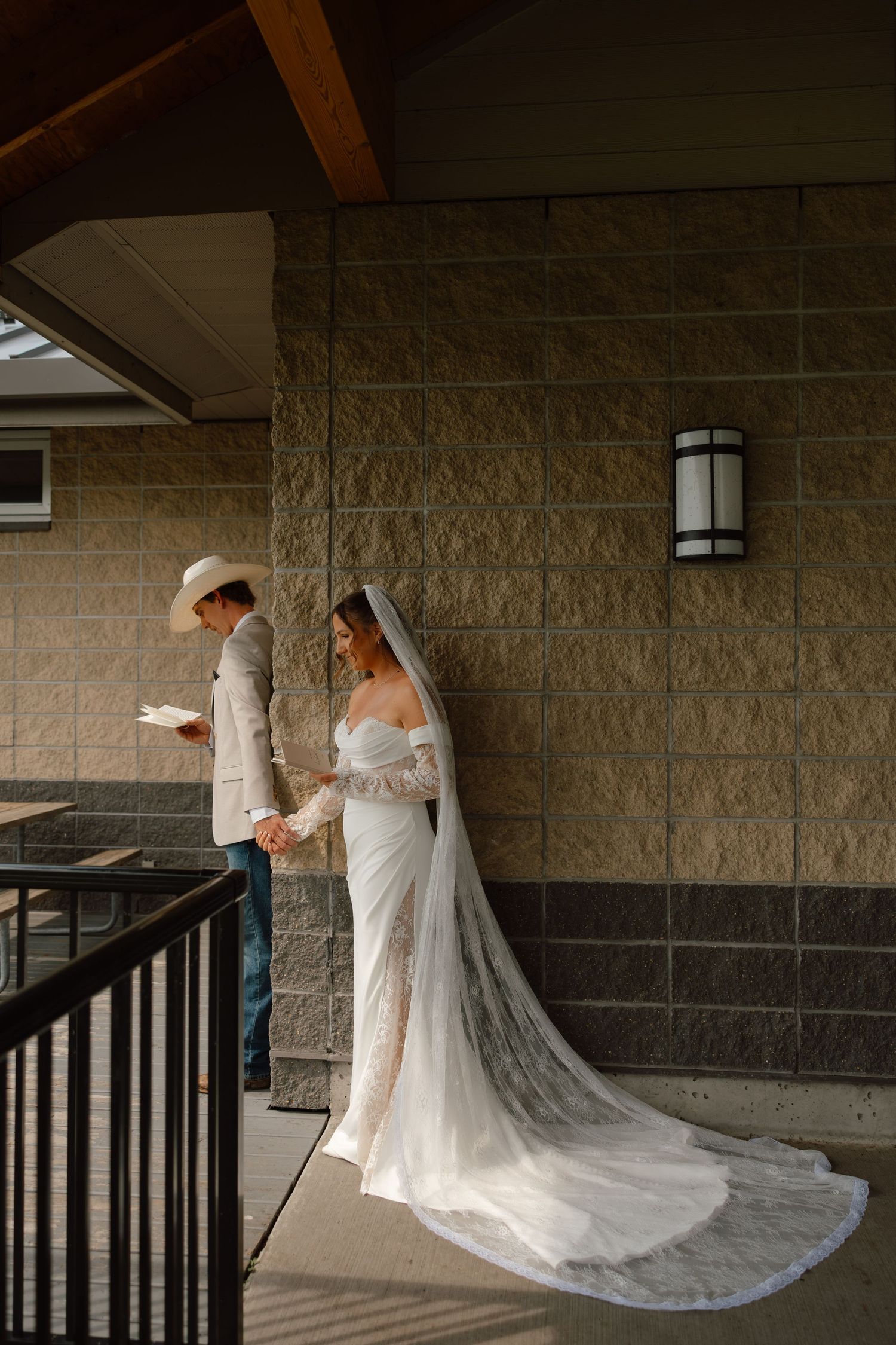 Bride in lace gown and veil holds hands with groom in cowboy hat reading vows on a rustic covered porch.