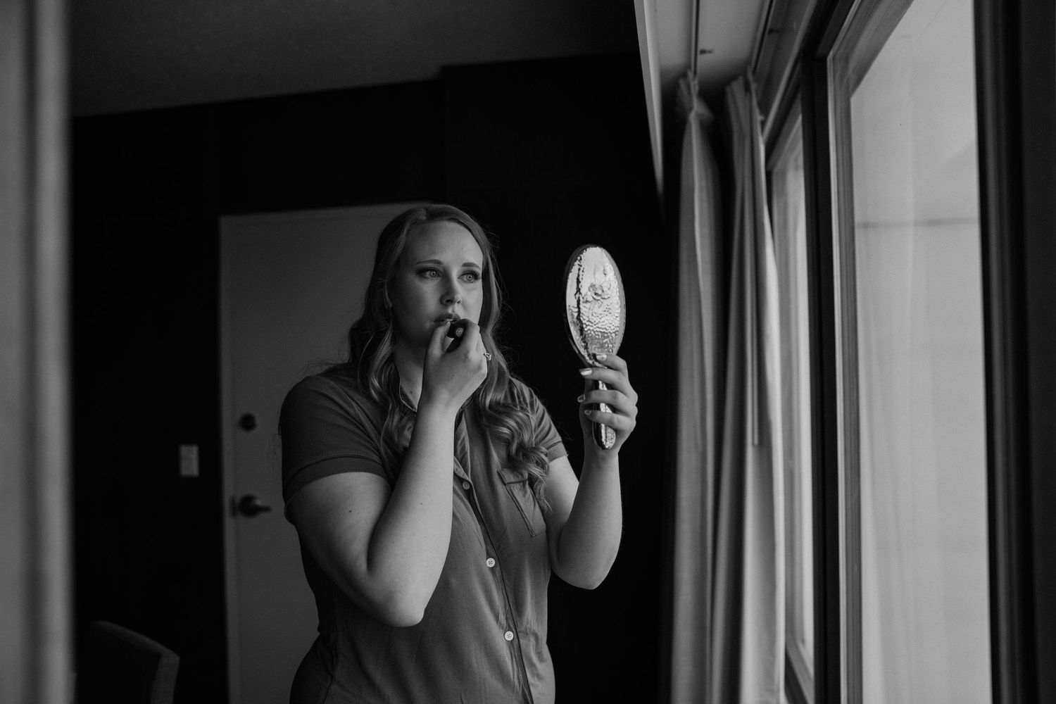 Woman applying lipstick while holding a small mirror in a black and white photo near a window.