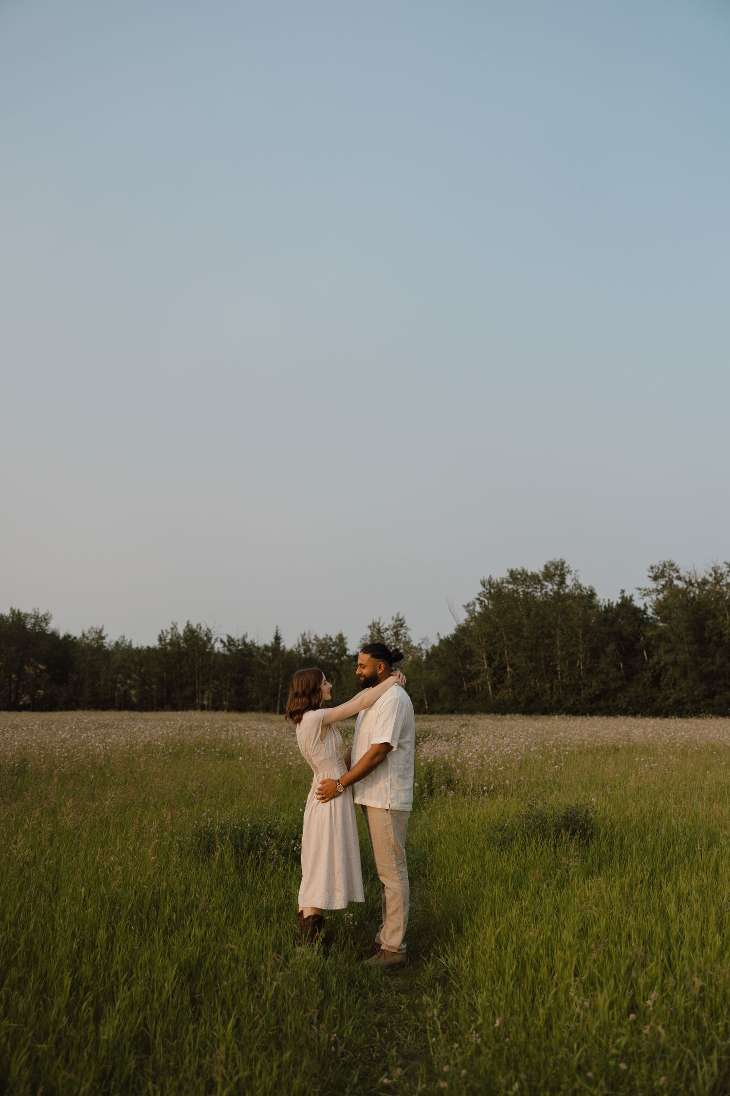 A couple embraces in a lush green field at dusk, wearing white outfits, with trees in the background.