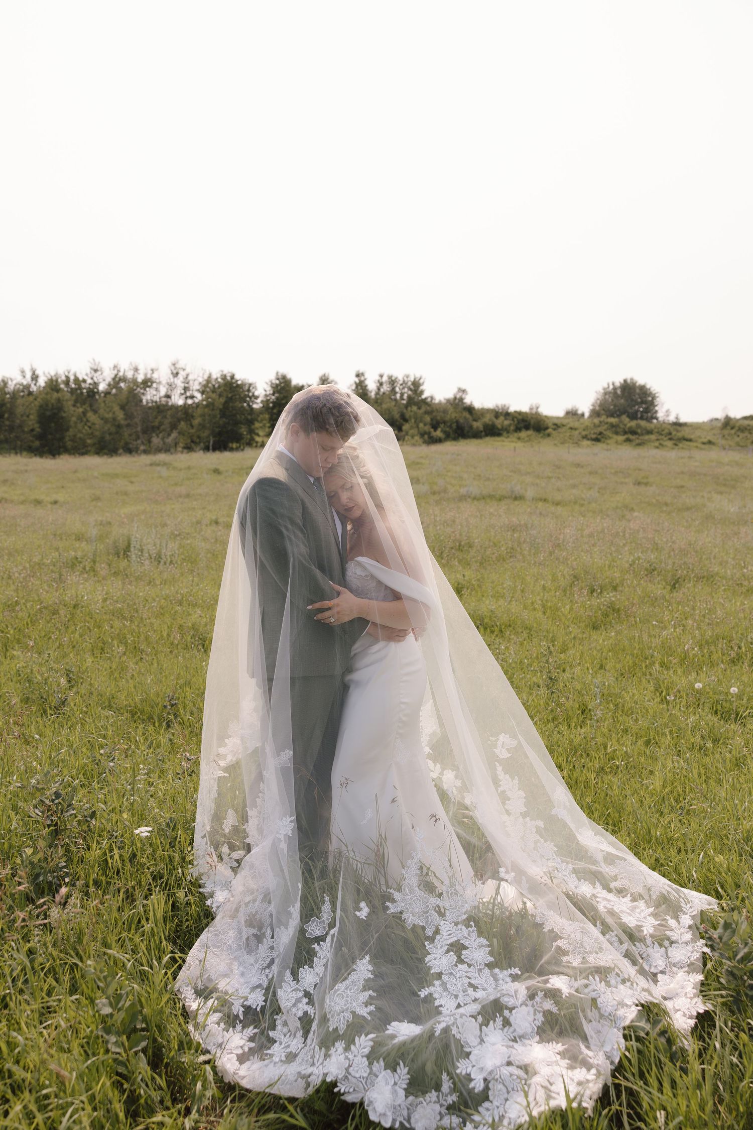 Bride and groom embrace under a flowing lace veil in a green meadow, sharing a romantic wedding moment outdoors.