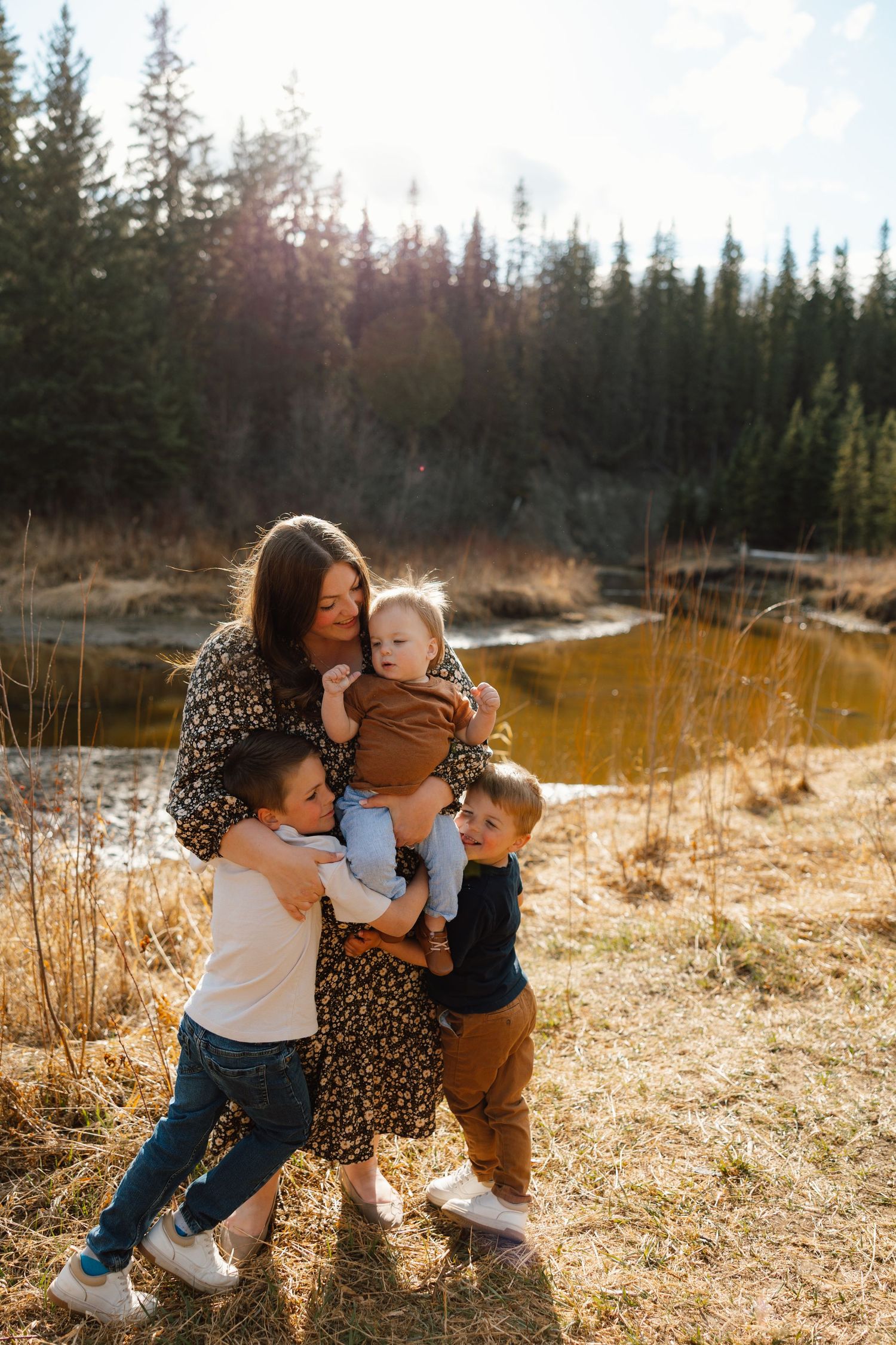 Mother holding baby while two young sons hug her near a sunlit forest pond in autumn.