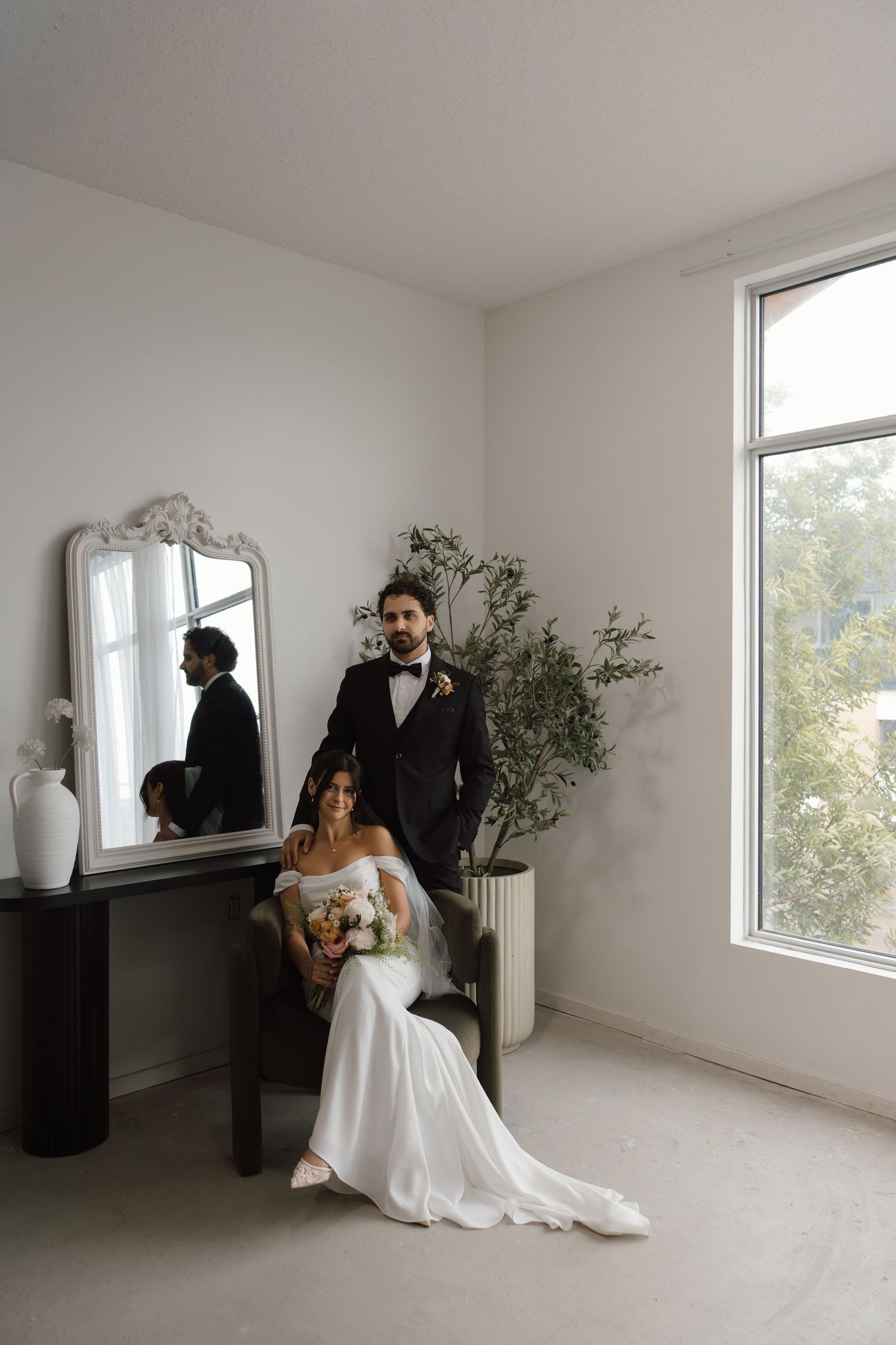 Bride seated in chair holding bouquet with groom standing behind her in tuxedo, elegant white room with mirror and window.