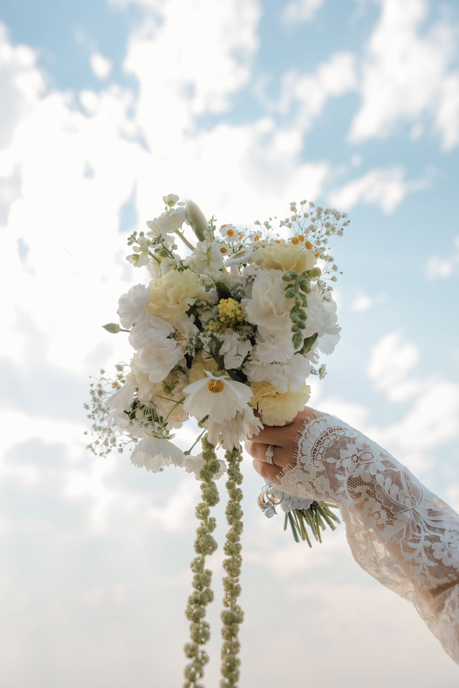 Bride in lace sleeve holding white wedding bouquet with daisies and greenery raised against a cloudy blue sky.