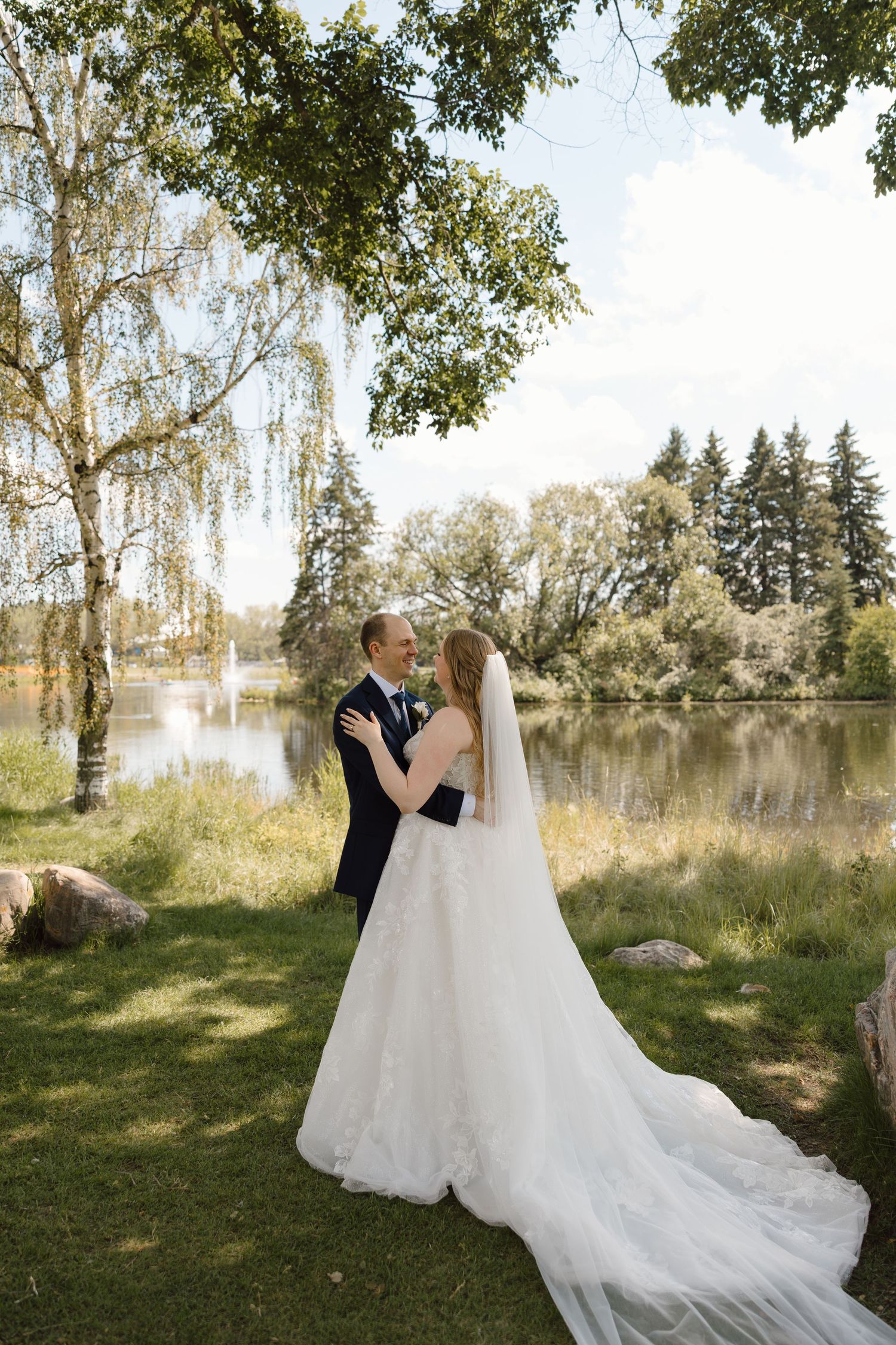 Bride and groom share a romantic moment by a serene lake surrounded by lush trees on their wedding day.