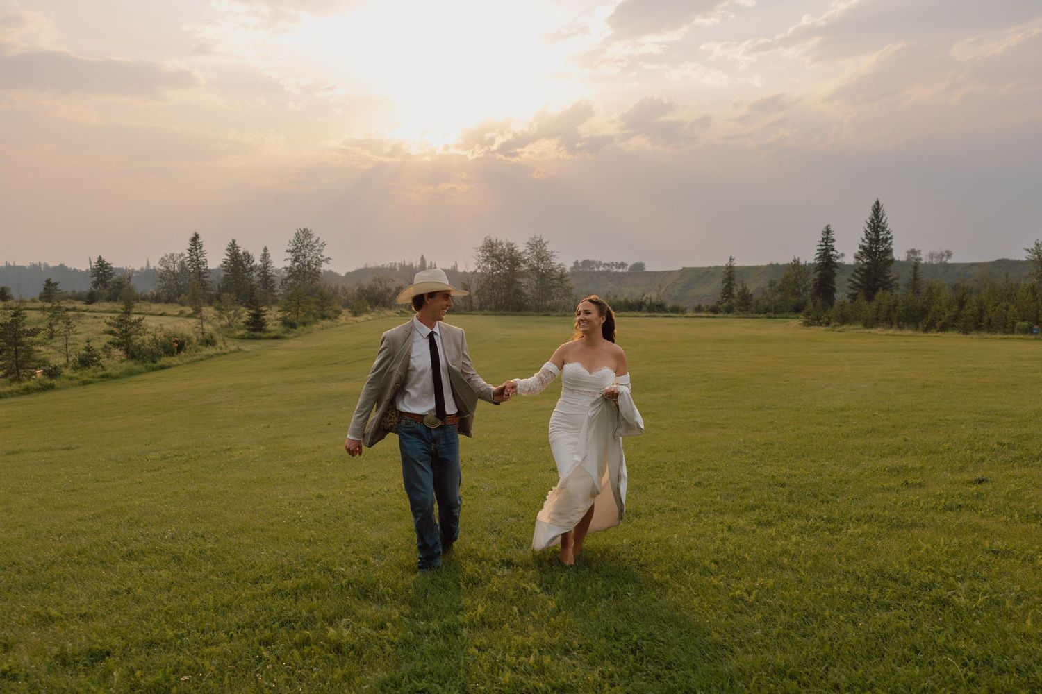 Bride and groom holding hands walking through green meadow at golden sunset, groom in cowboy hat, bride in white gown.