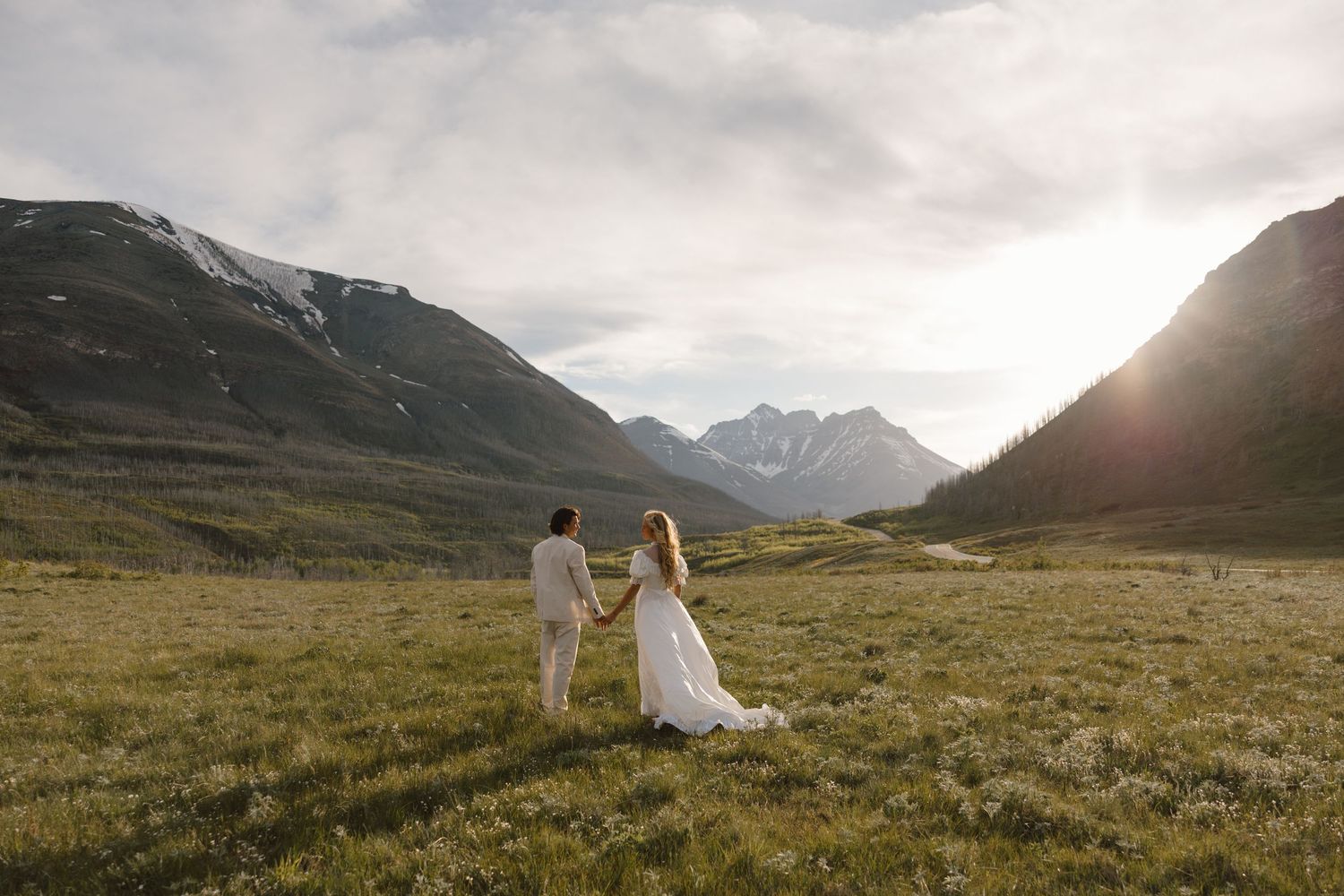 Couple in wedding attire holding hands in a lush mountain valley with sunlight streaming through peaks.