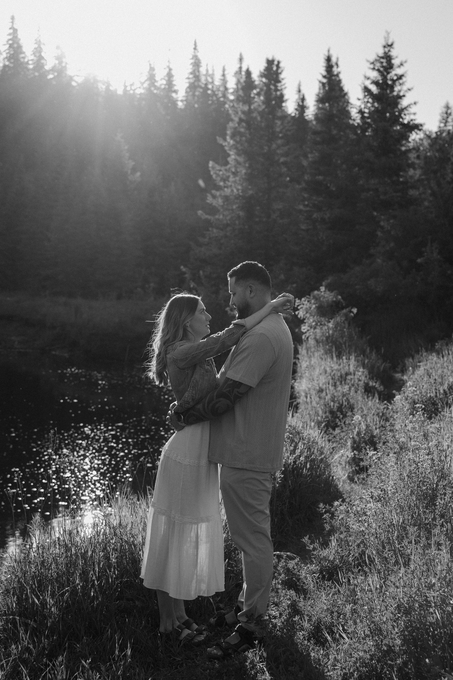 Black and white photo of a couple embracing near a mountain lake surrounded by pine trees in golden sunlight.