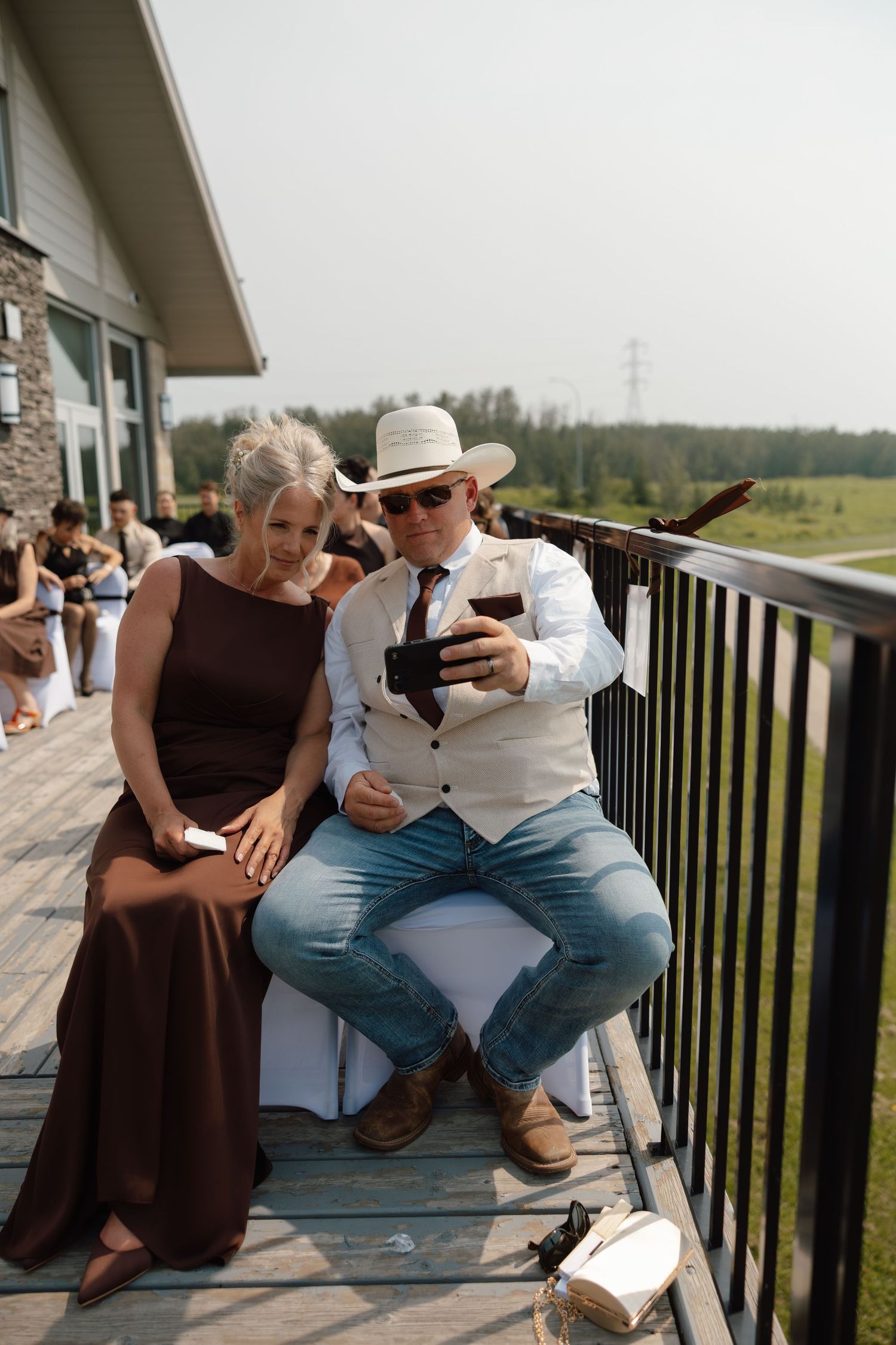 Man in cowboy hat and woman in brown gown share a selfie moment on a wooden deck at an outdoor wedding.