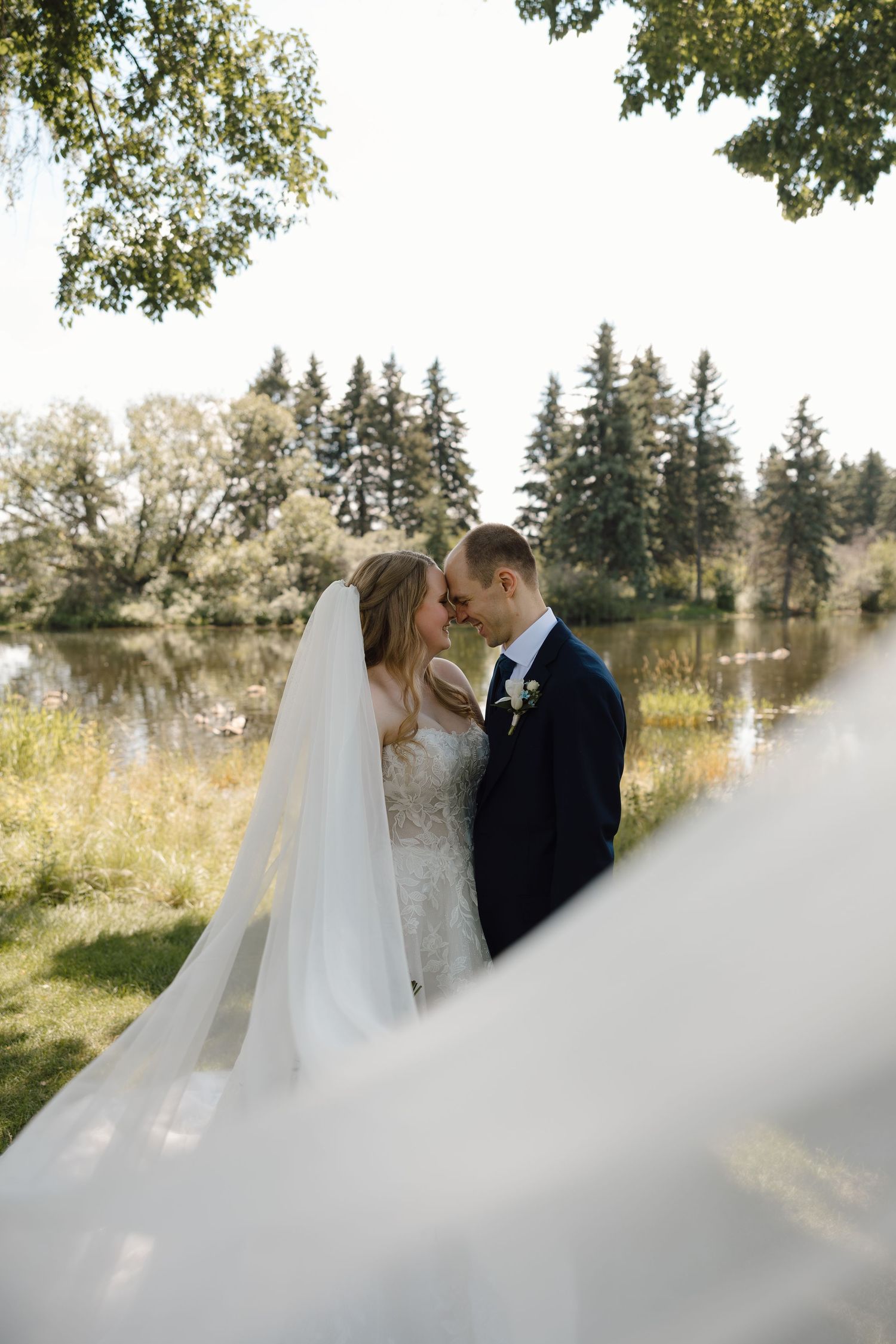 Bride and groom share a romantic moment by a sunlit lake, her veil flowing in the breeze, surrounded by lush trees.