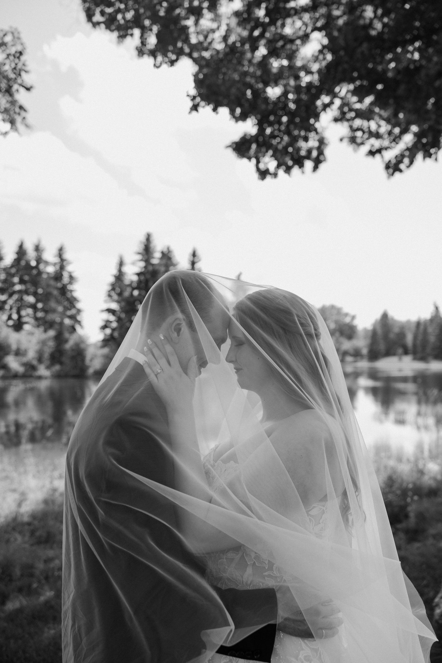 Black and white wedding photo of bride and groom sharing intimate moment under veil by a lake.