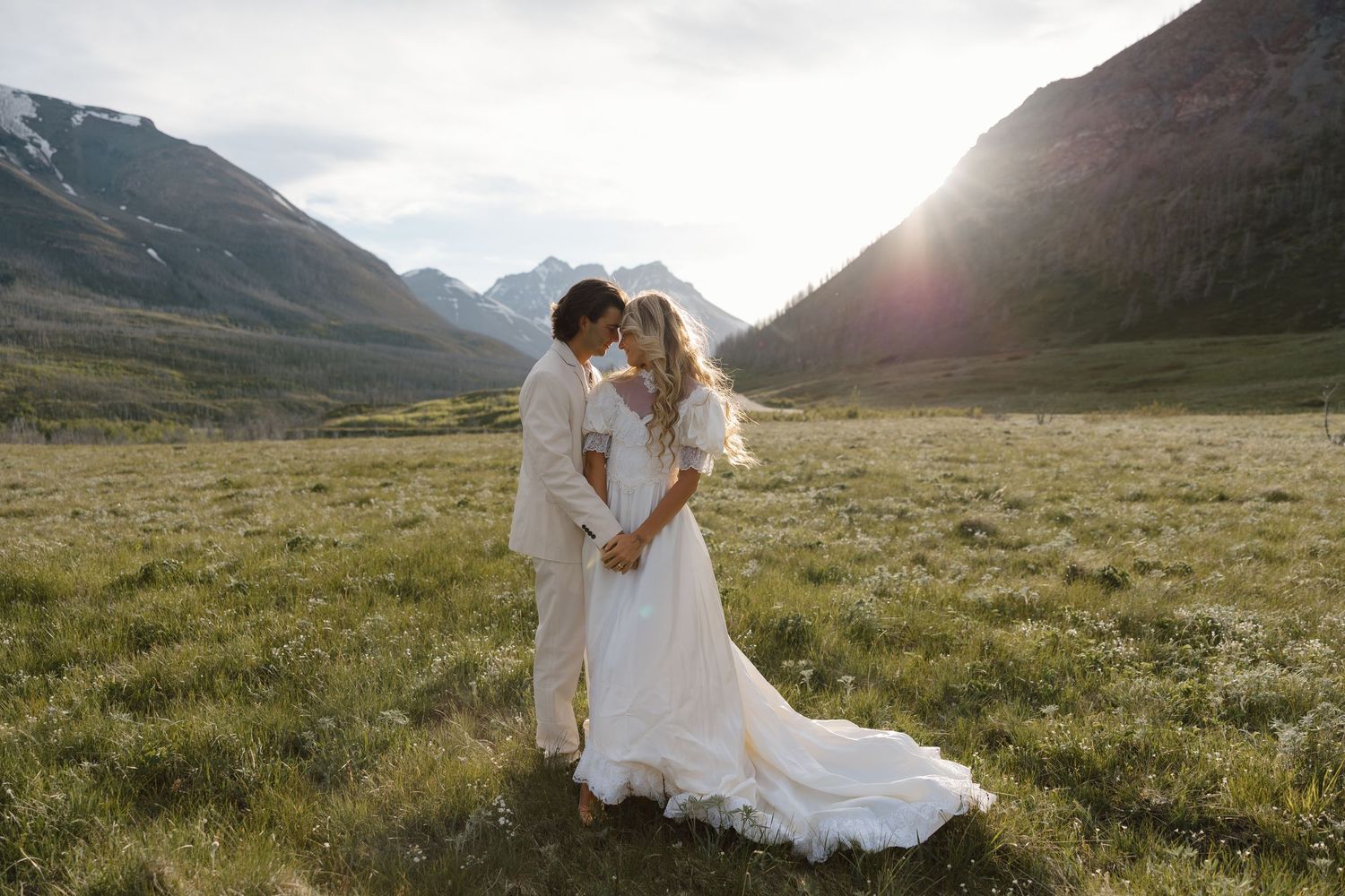 A bride and groom share a romantic moment in a lush mountain meadow at sunset, surrounded by majestic peaks.