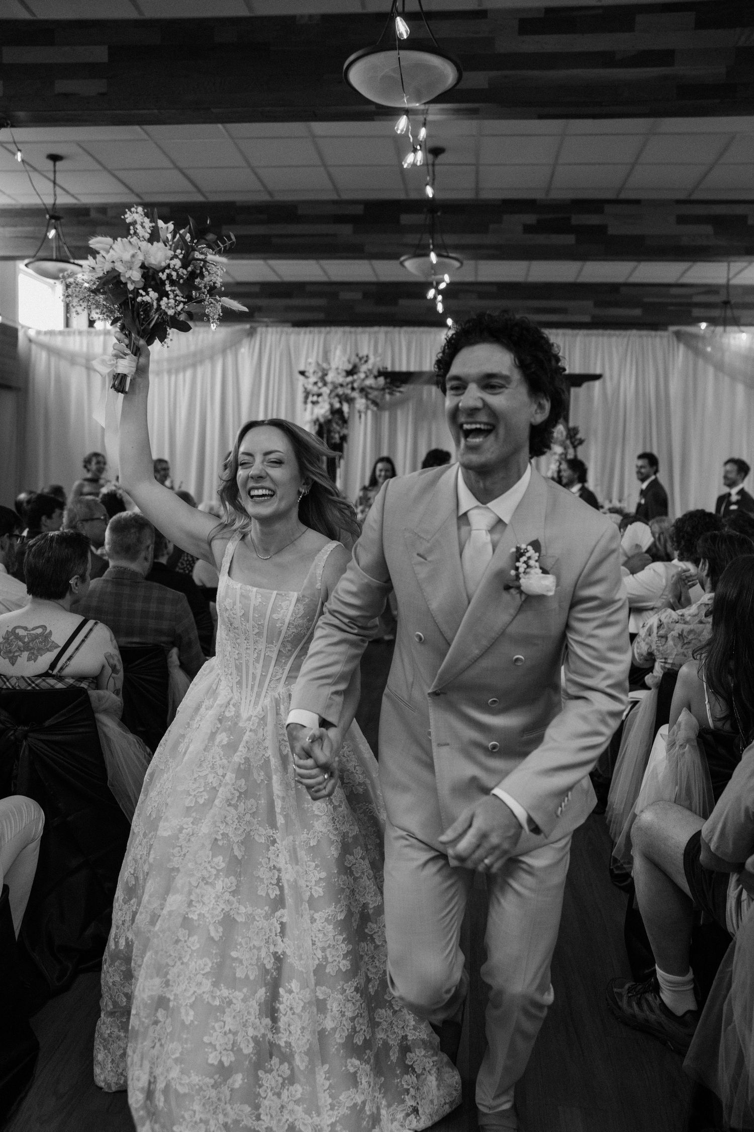 Black and white photo of joyful newlyweds walking down the aisle, bride holding bouquet aloft, groom in suit, guests cheering.