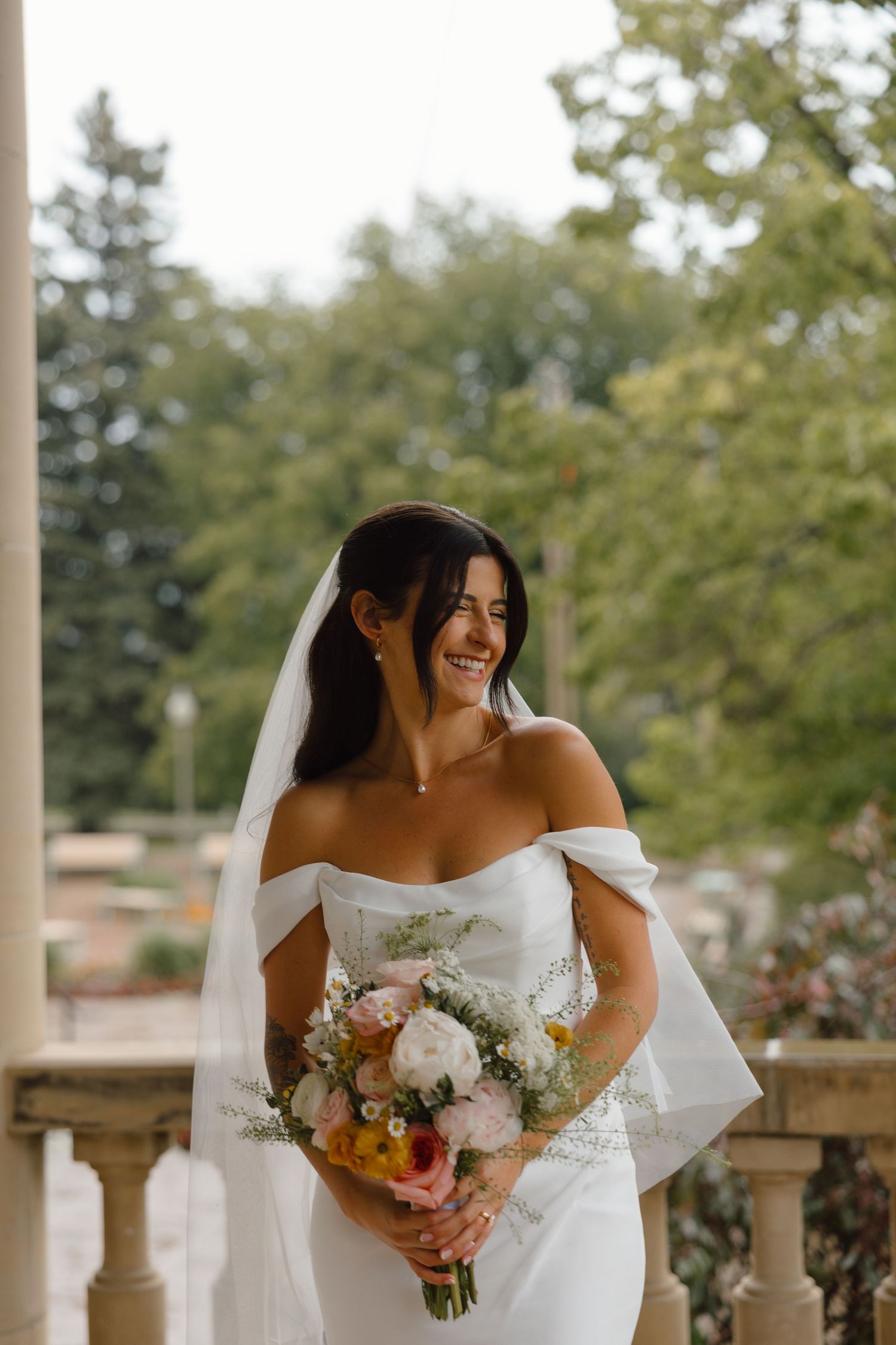 Smiling bride in off-shoulder white gown and veil holds colorful bouquet on elegant columned terrace with lush trees.