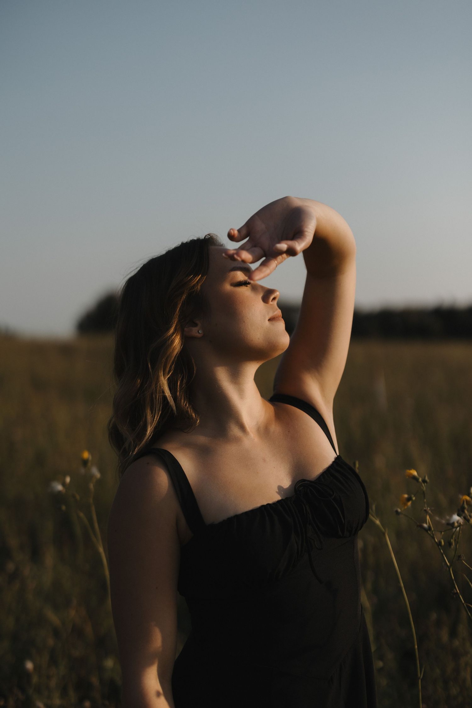 Woman in black dress standing in a golden field at sunset with hand raised to her forehead.