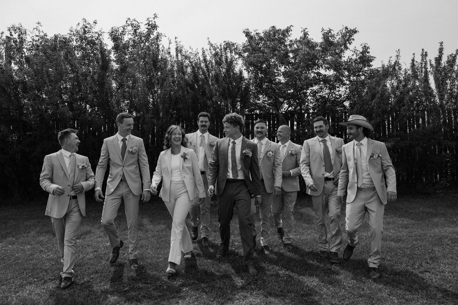 Black and white photo of a wedding party in light suits walking together outdoors near tall hedges.