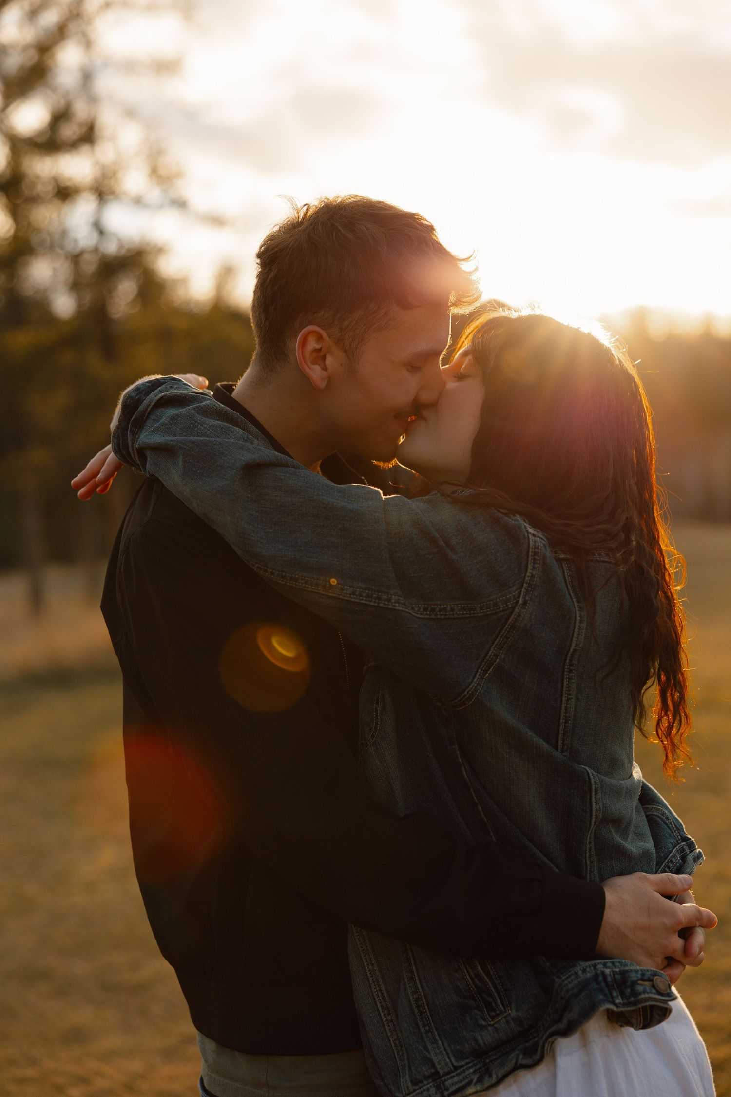 Couple embracing and kissing at golden sunset in an open field with warm lens flare.