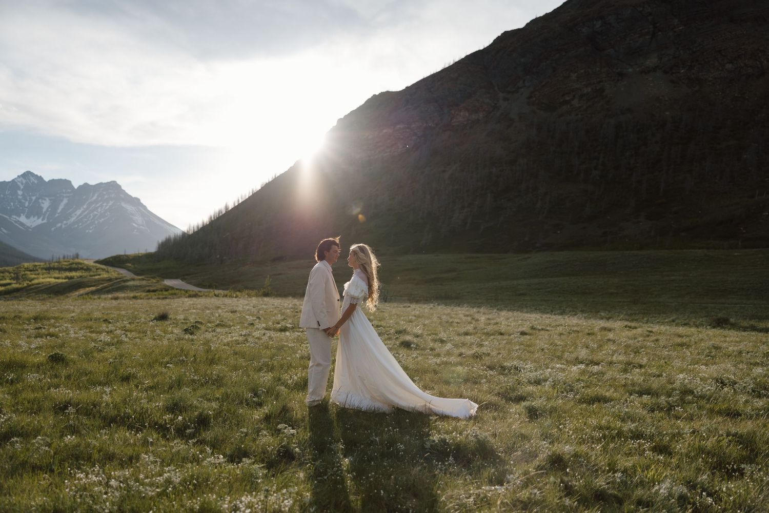 Bride and groom embrace in sunlit alpine meadow with mountain backdrop during golden hour outdoor wedding.