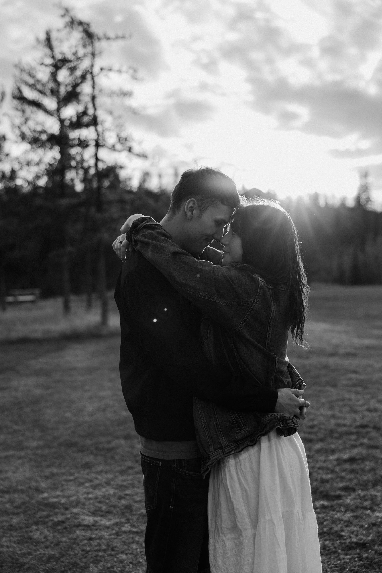 Black and white romantic photo of couple embracing at sunset in a forest clearing with dramatic backlight. Taken in Edmonton, Alberta, Canad